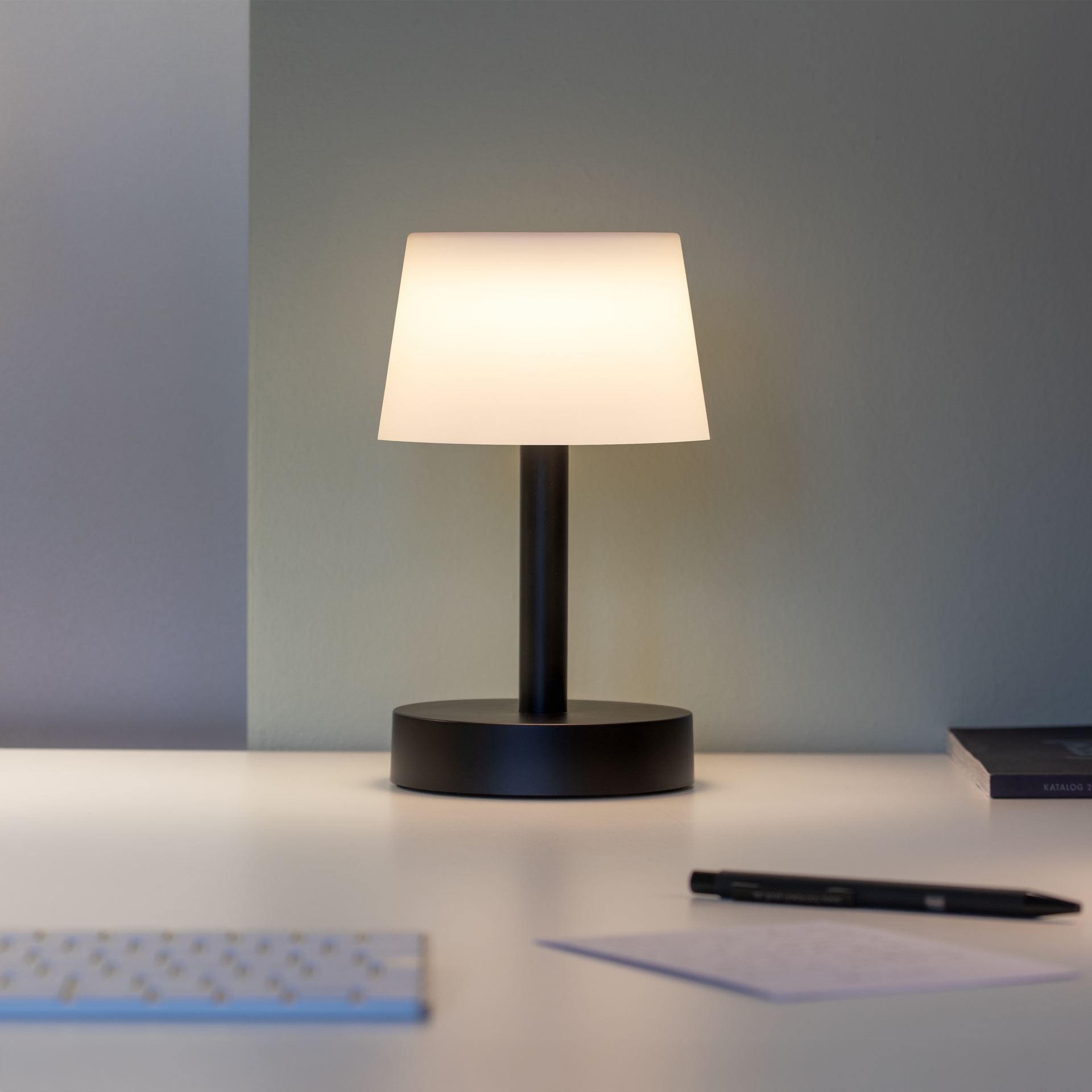 La lámpara de mesa «Fritz», con pie negro y pantalla clara, ilumina una mesa blanca. Junto a ella hay un bolígrafo, papel y un folleto.