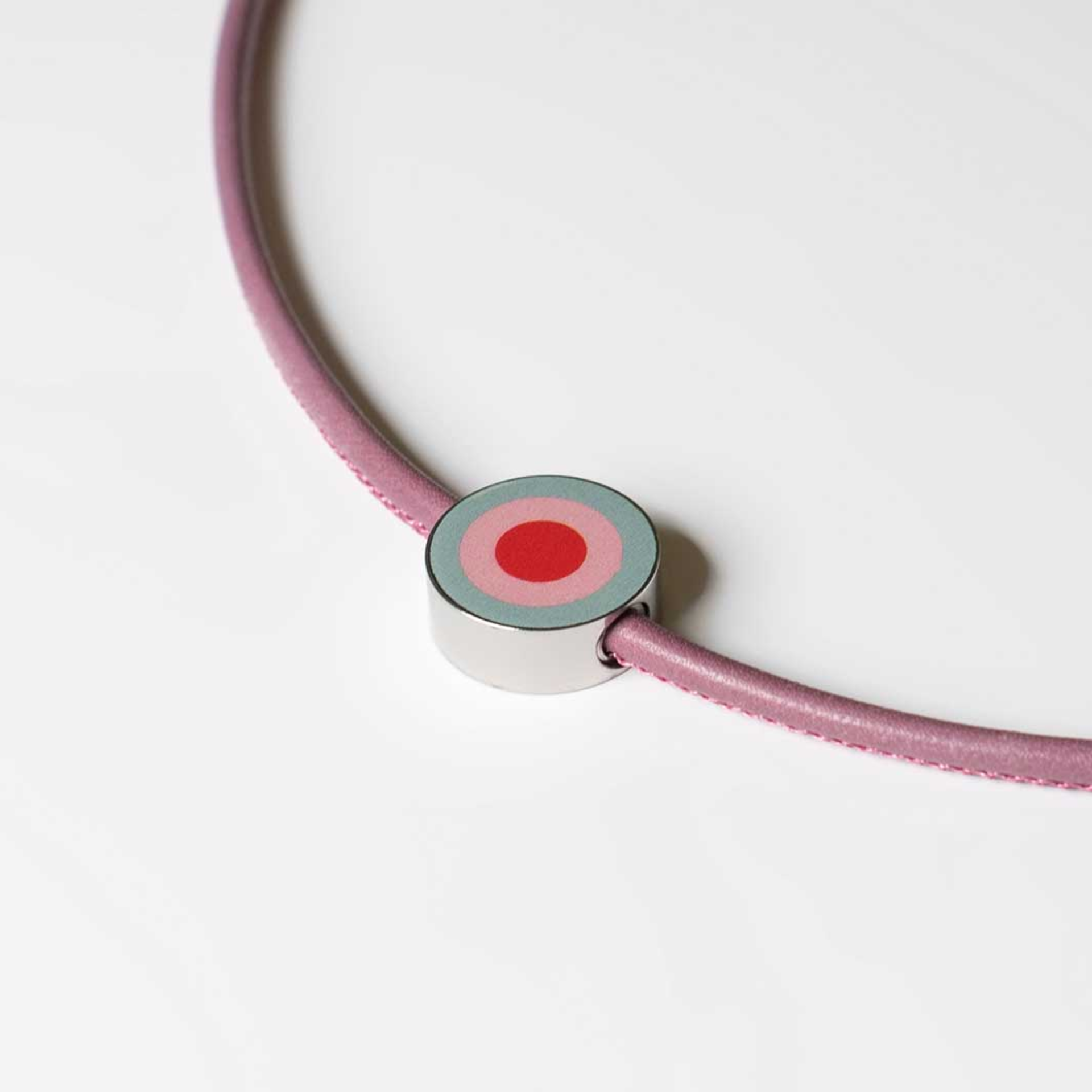 Primo piano di un ciondolo da gioielleria di colore lilla su un nastro. Il ciondolo presenta un motivo di cerchi in rosso, rosa e azzurro. Il ciondolo è in acciaio.