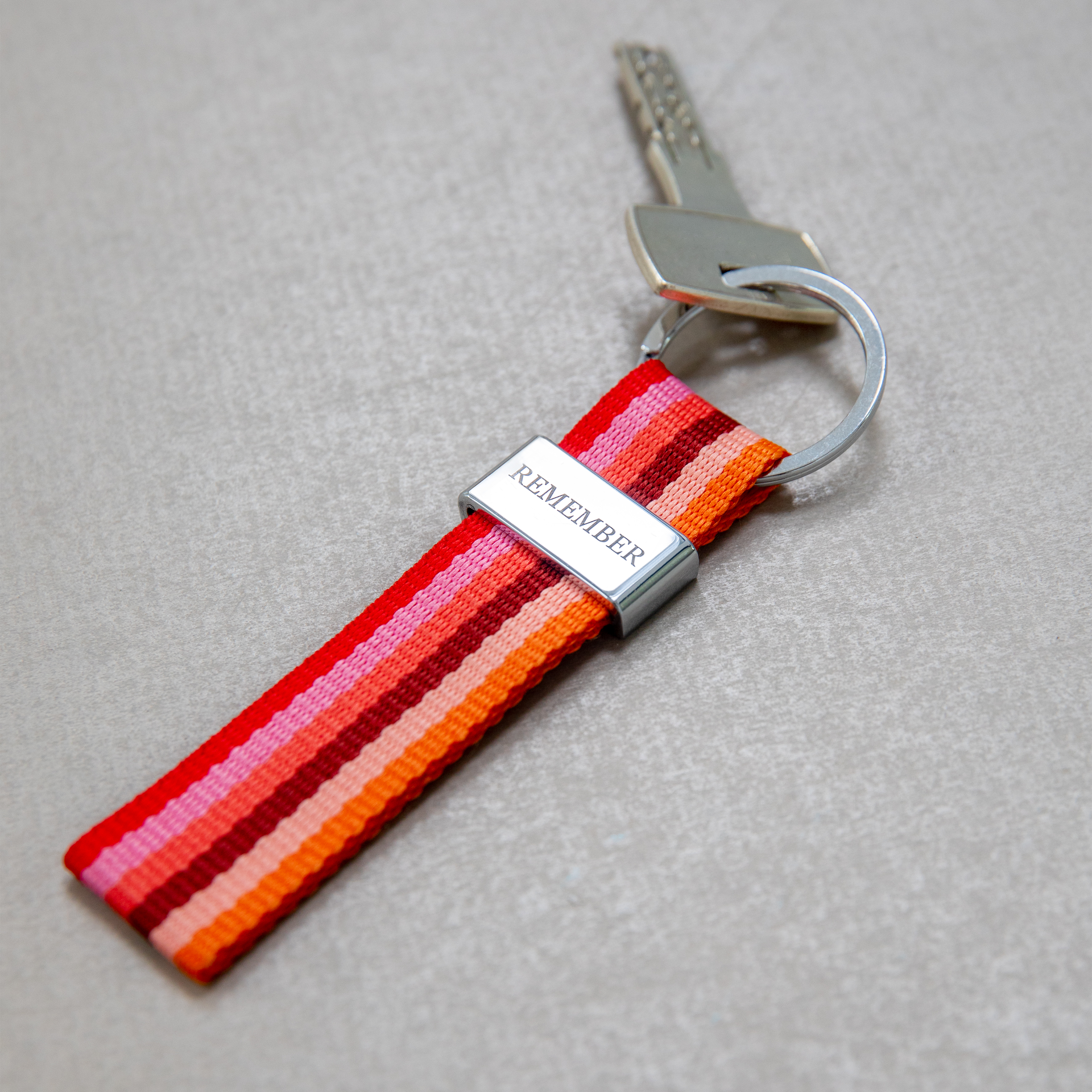 Schlüsselband mit lebhaftem, vertikalem Streifenmuster in Rot-, Pink-, Orange- und Brauntönen. Oben ist eine glänzende, silberne Metallplakette mit eingraviertem „REMEMBER“ befestigt. Mit robustem Schlüsselring.