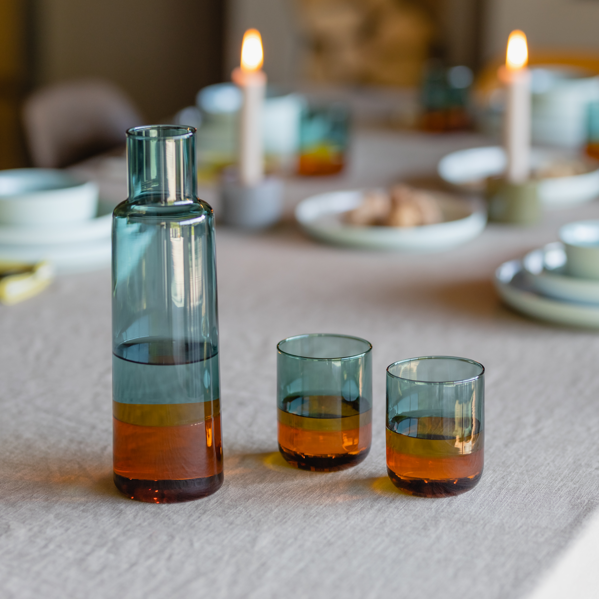 Two amber-coloured drinking glasses and a matching carafe stand on a laid table. Crockery and candles can be seen in the background.
