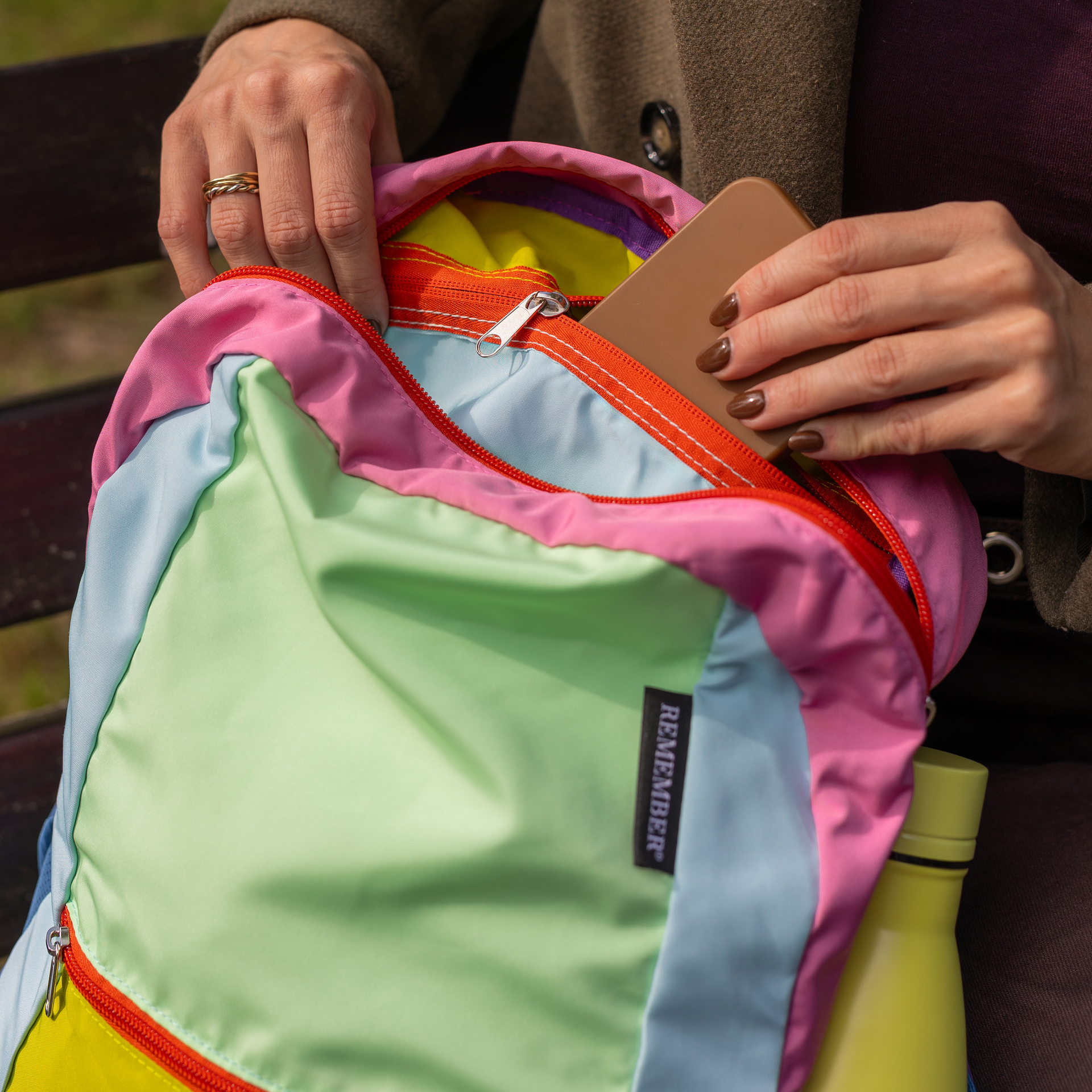 Hände mit braunem Nagellack stecken ein Smartphone in das Hauptfach eines bunten, faltbaren Polyester-Rucksacks. Hellgrün, Hellblau, Rosa, rote Reißverschlüsse, schwarzes "REMEMBER" Etikett. Gelbe Flasche in Netztasche.