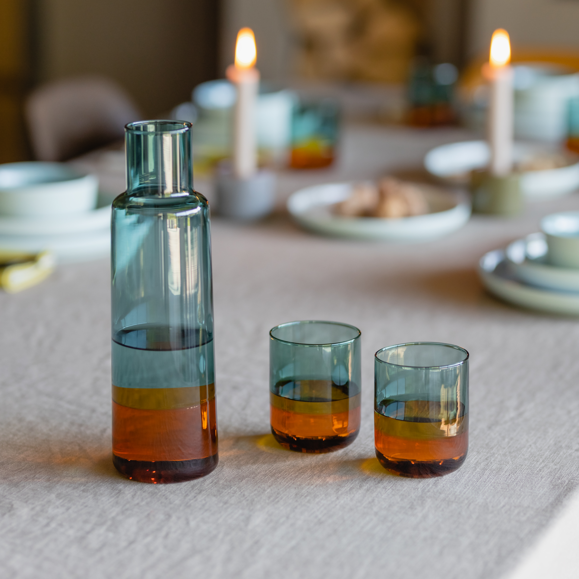 Una caraffa di vetro "ambrato" e due bicchieri su un tavolo. Gli oggetti di vetro sono parzialmente riempiti di un liquido ambrato. Sullo sfondo si vede una tavola imbandita con candele accese.