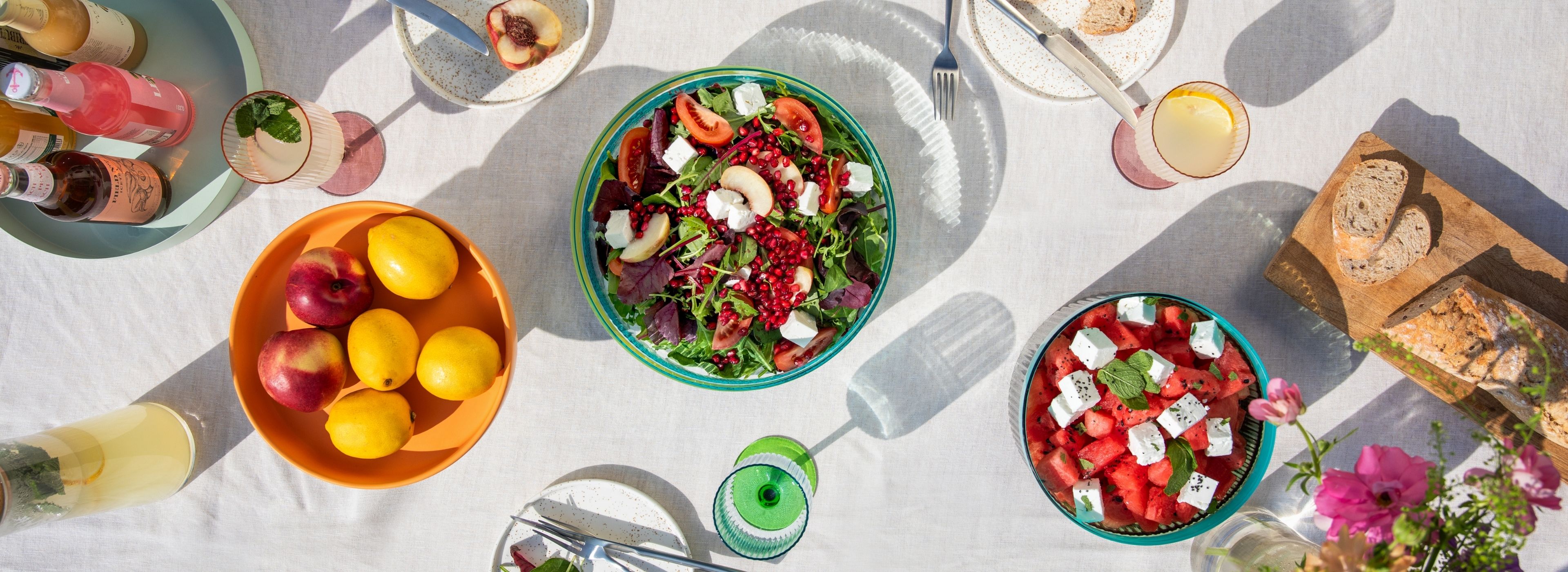 Ein Sommertisch im Freien, reich gedeckt mit Speisen und Getränken. Zwei Salate: Blattsalat mit Pfirsich, Feta, Granatapfelkernen; Wassermelone mit Feta, Minze. Dazu Zitronen, Pfirsiche, Flaschen und Brot.