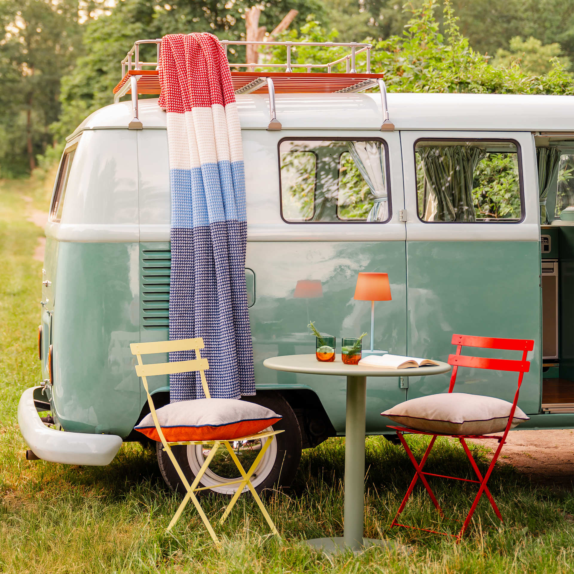 Una furgoneta VW de color menta con una manta «Malie» de colores rojo, blanco y azul en el portaequipajes del techo. Delante, una mesa con una lámpara y dos sillas con cojines en plena naturaleza.