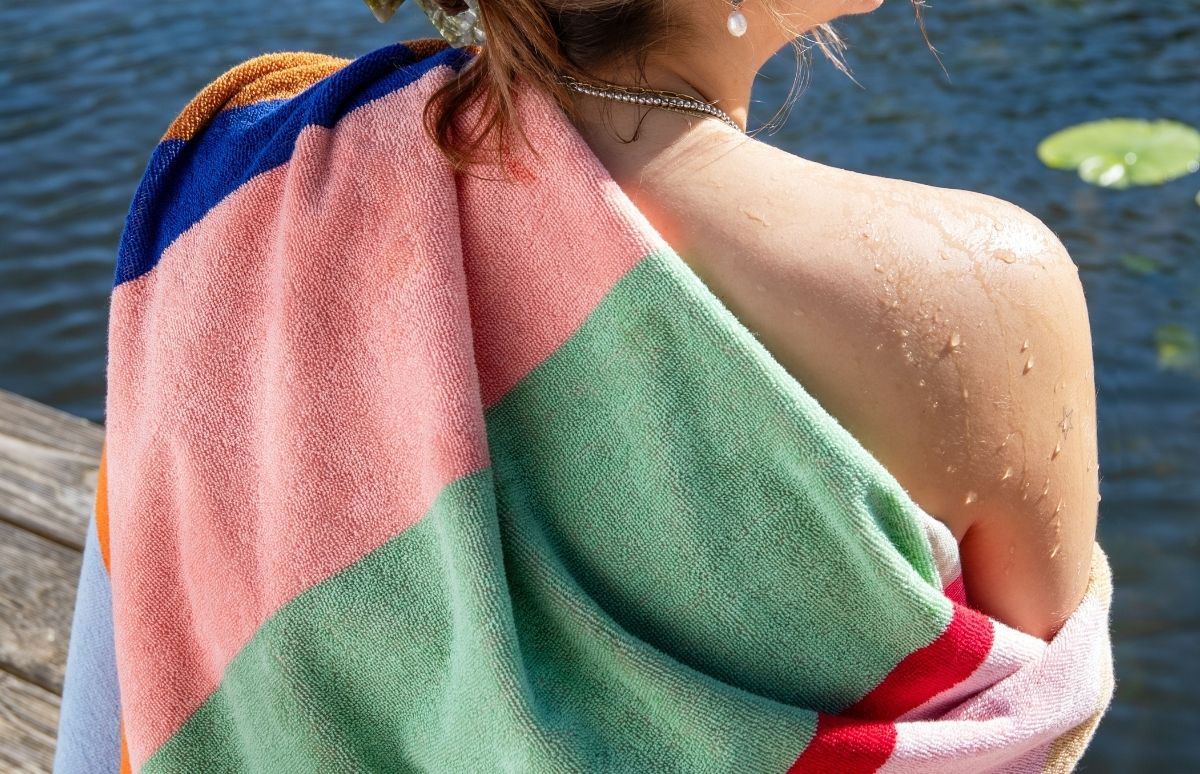 Eine Person sitzt nach dem Schwimmen auf einem Holzsteg am Wasser, eingehüllt in ein bunt gestreiftes Handtuch in Rosa, Grün und Blau. Wassertropfen glänzen auf Schulter und Rücken; im Hintergrund treibt ein Seerosenblatt.