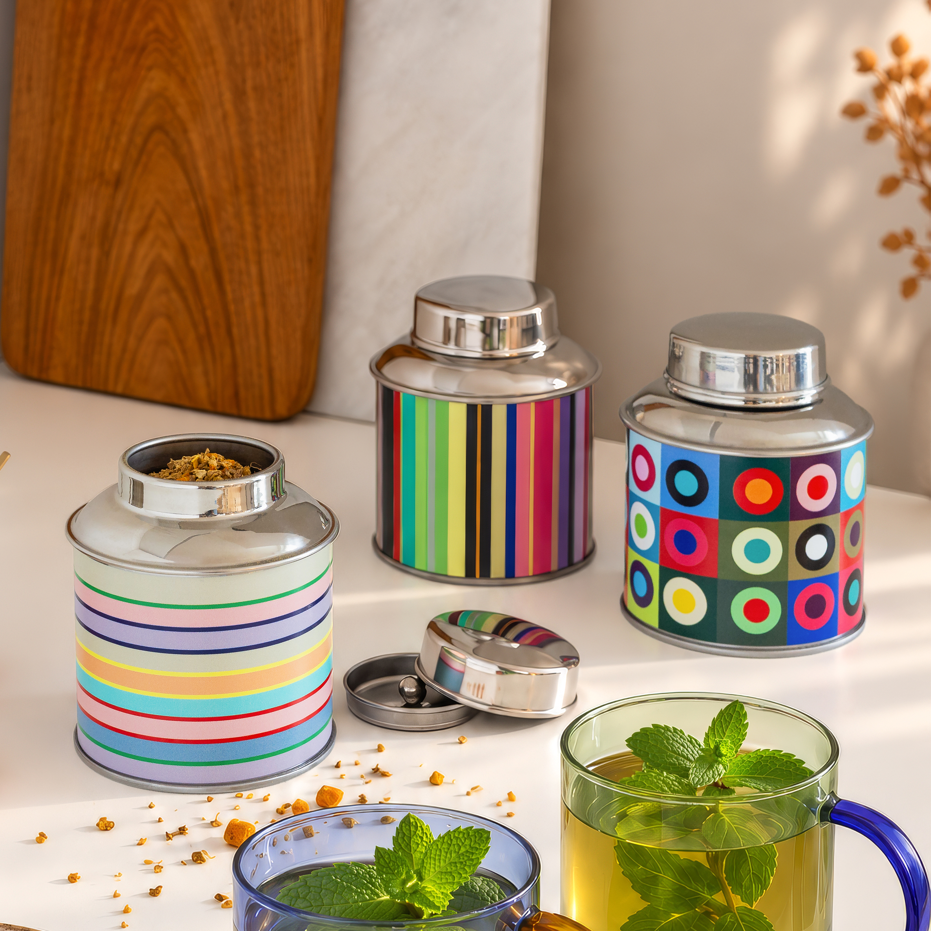 Trois boîtes à thé colorées en acier inoxydable sont sur une table blanche. Celle de gauche, à rayures, est ouverte avec du thé en vrac; quelques feuilles sont éparpillées. Devant, deux verres de thé à la menthe.