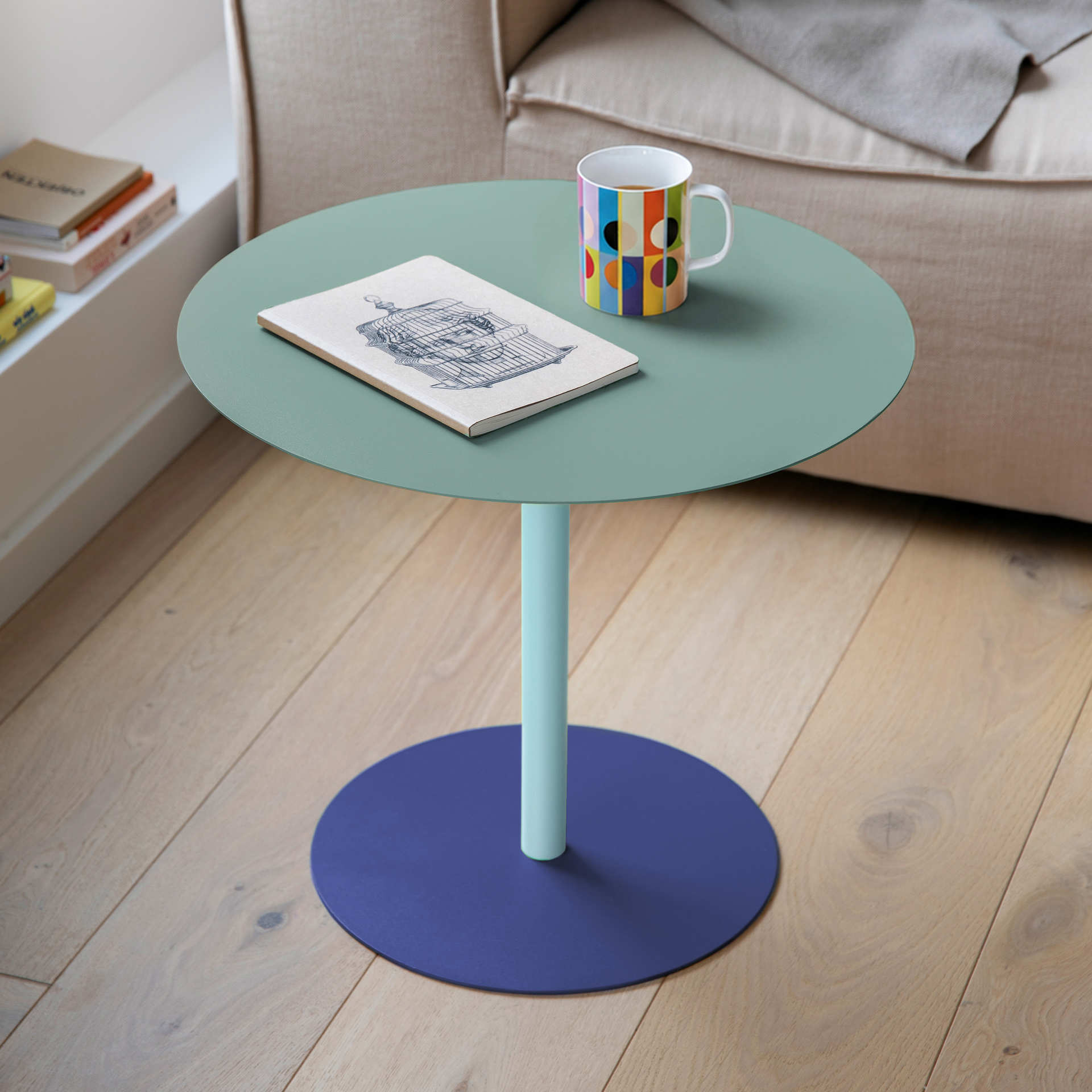 Ronde bijzettafel Rondo met mintgroen tafelblad, lichtblauwe kolompoot en donkerblauwe ronde voet op een houten vloer. Op het tafelblad liggen een notitieboekje met een tekening van een vogelkooi en een kleurrijke kop. Lichte bank op de achtergrond.