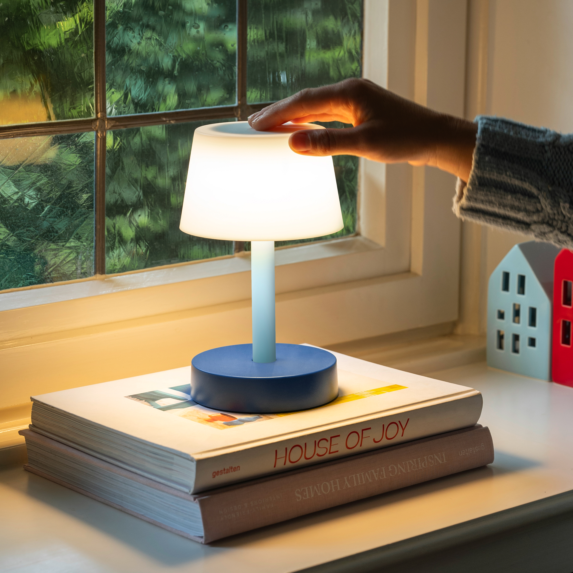 De Fritz 'sky' tafellamp op boeken. Een hand raakt de verlichte kap aan. Op de achtergrond zijn een raam en decoratieve huizen te zien.