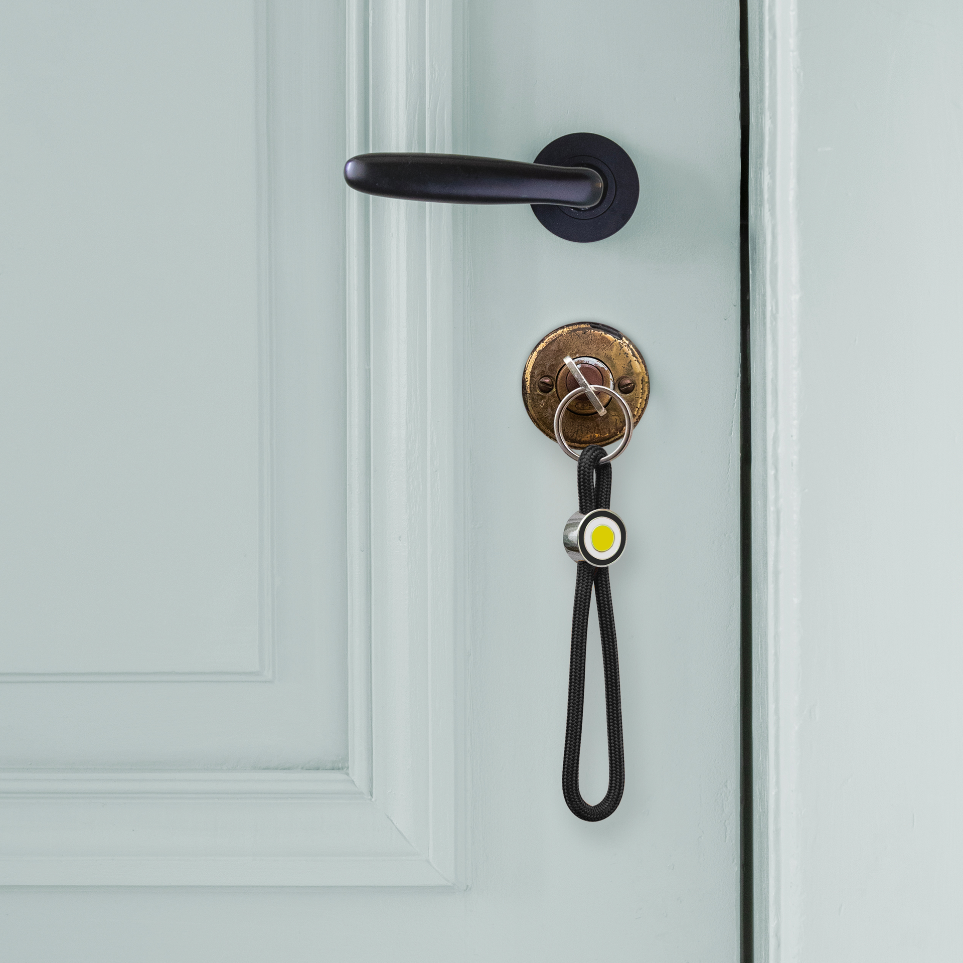 Schwarzer Schlüsselanhänger aus geflochtenem Seil in Schlaufenform. In der Mitte hält ein glänzender Metallring das Seil zusammen, mit schwarzer Umrandung und leuchtend gelbem Kreis im Ringo-Design. Unten ein silberner Schlüsselring.