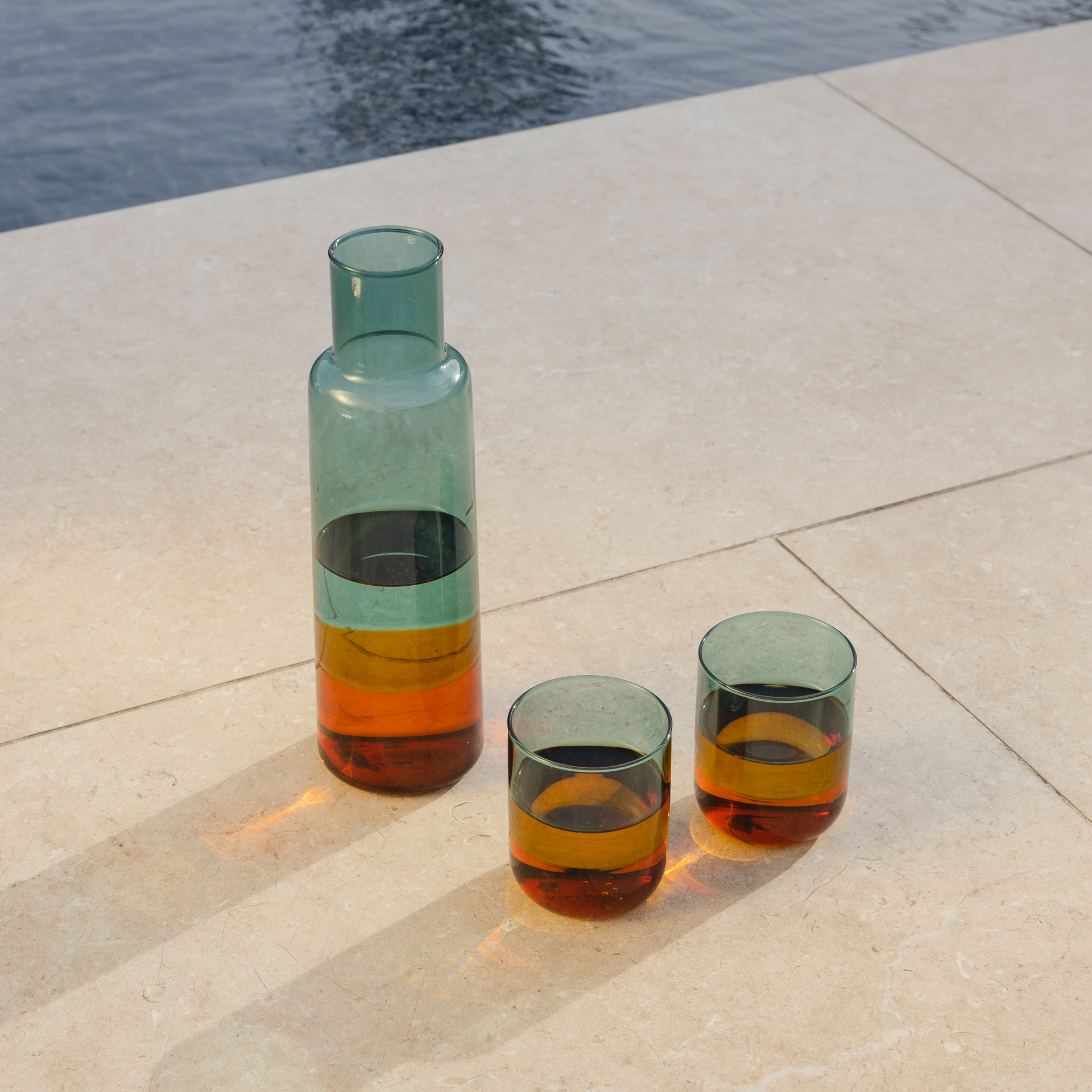 Una caraffa di vetro alta e stretta e due bicchieri poggiano su una superficie di pietra chiara. I recipienti sono pieni di un liquido ambrato e riflettono la luce. Sullo sfondo è visibile dell'acqua.