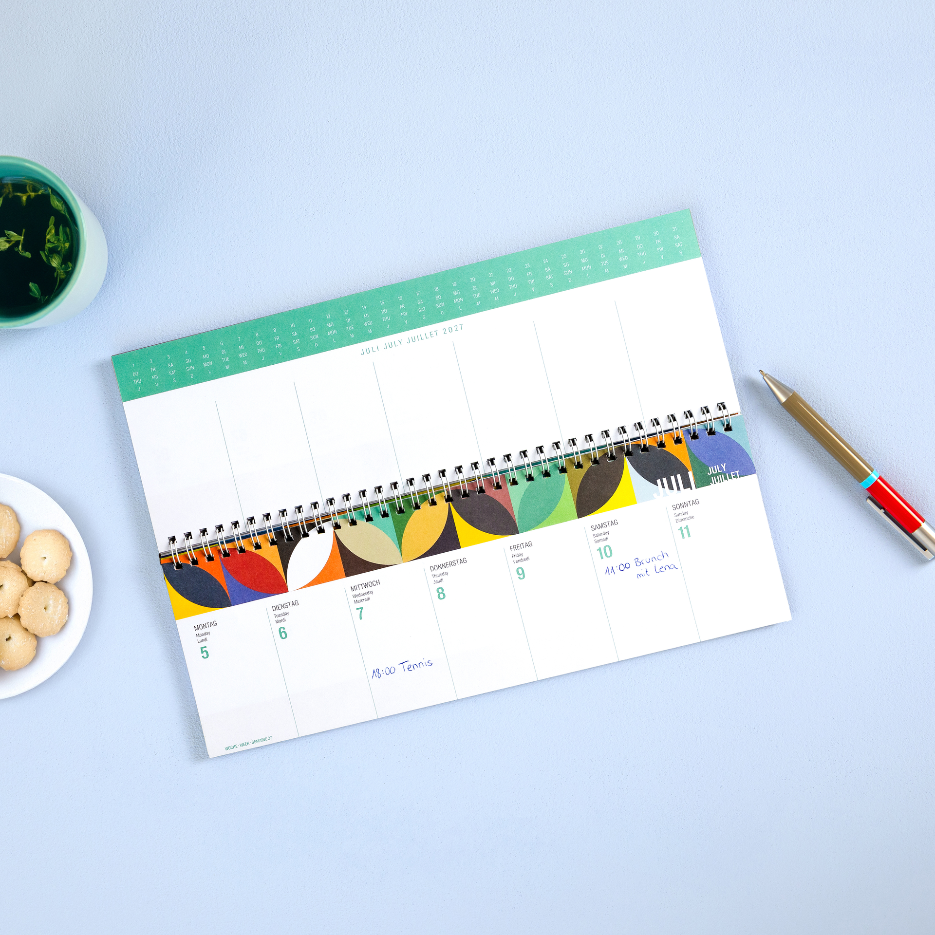 Colourful weekly planner 2027 with top-bound spiral binding, ideal for desk-based scheduling. Front cover: vibrant abstract pattern of geometric shapes in blue, green, red and yellow. White lettering: ‘REMEMBER ’ and ‘WEEKLY PLANNER 2027’.