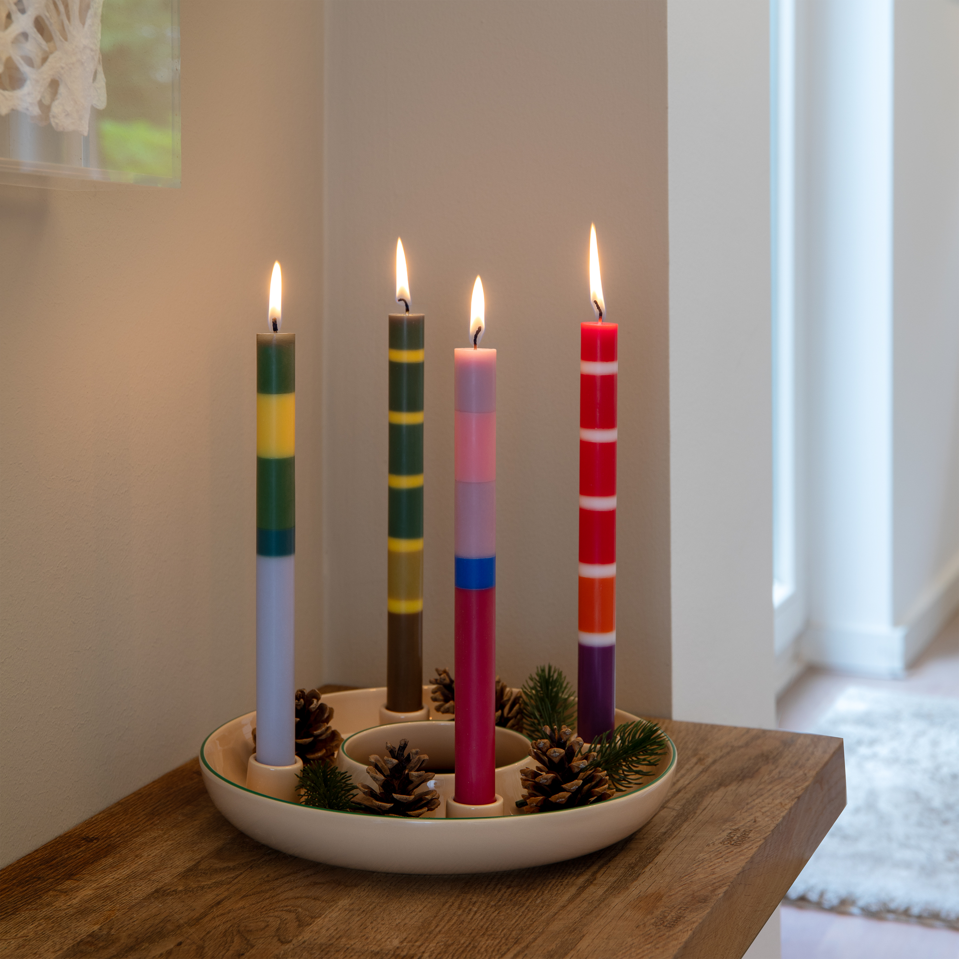 Sobre una mesa de madera hay un portavelas de color beige con el borde verde. En él arden cuatro velas cilíndricas con rayas de colores: verde, amarillo, azul, rosa y rojo. Entre ellas hay piñas y ramitas verdes.