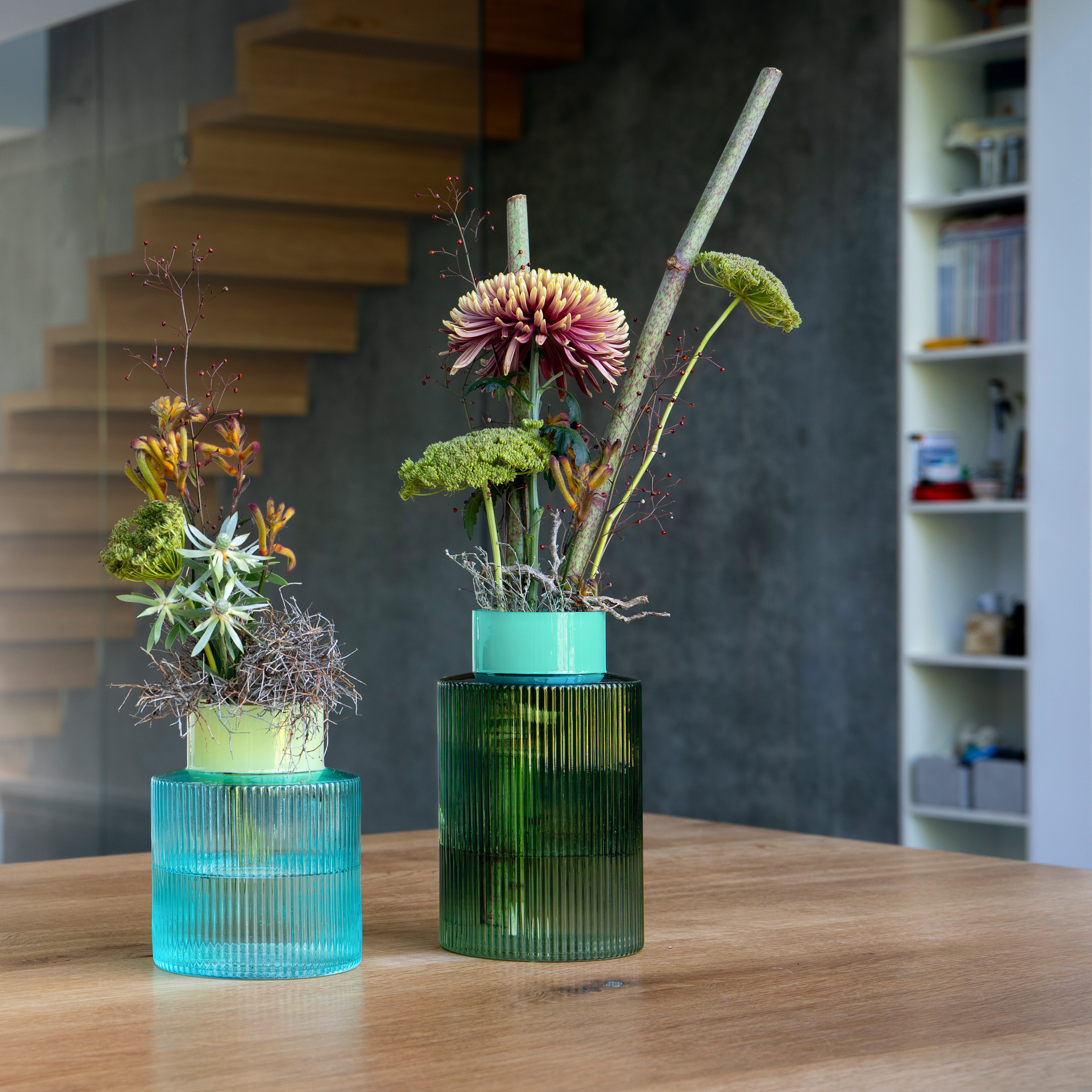 Two fluted glass vases, one green, one blue, with floral decorations on a wooden table. Staircase and shelf in the background.