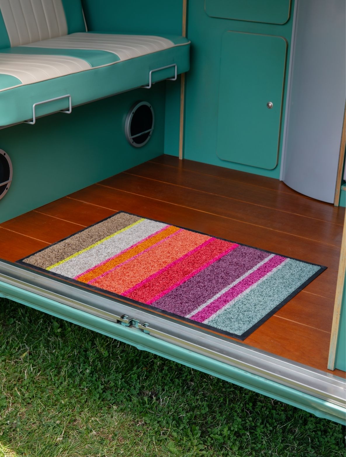 Ein bunter, gestreifter Fußabtreter liegt auf dem holzfarbenen Boden eines teal-farbenen Campervans. Im Hintergrund ist eine Sitzbank mit cremeweißen und teal-farbenen Polstern. Grünes Gras am Eingang deutet auf eine offene Tür hin.