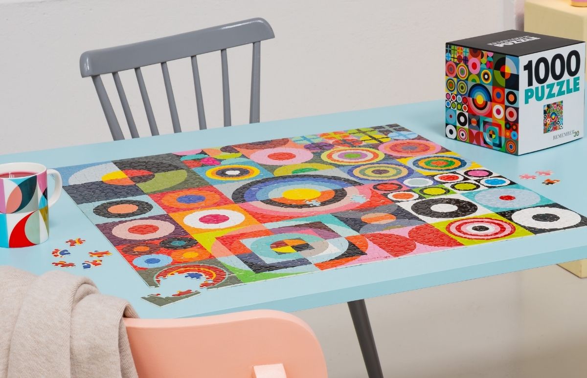Farbenfrohes, geometrisches Puzzle auf hellblauem Tisch mit abstraktem Muster aus bunten Kreisen und Quadraten. Daneben lose Teile. Links Tasse mit rotem Getränk, rechts Puzzlebox. Hinten grauer Stuhl, vorn beiger Schal.