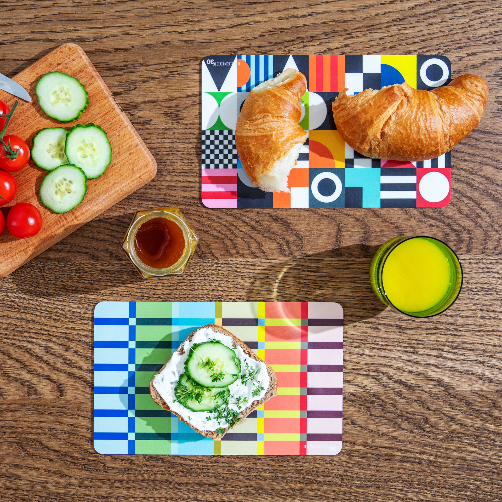 Draufsicht auf einen Frühstückstisch mit Croissants, Brot mit Frischkäse und Gurke, Tomaten, Honig und Saft auf bunten Tischsets.