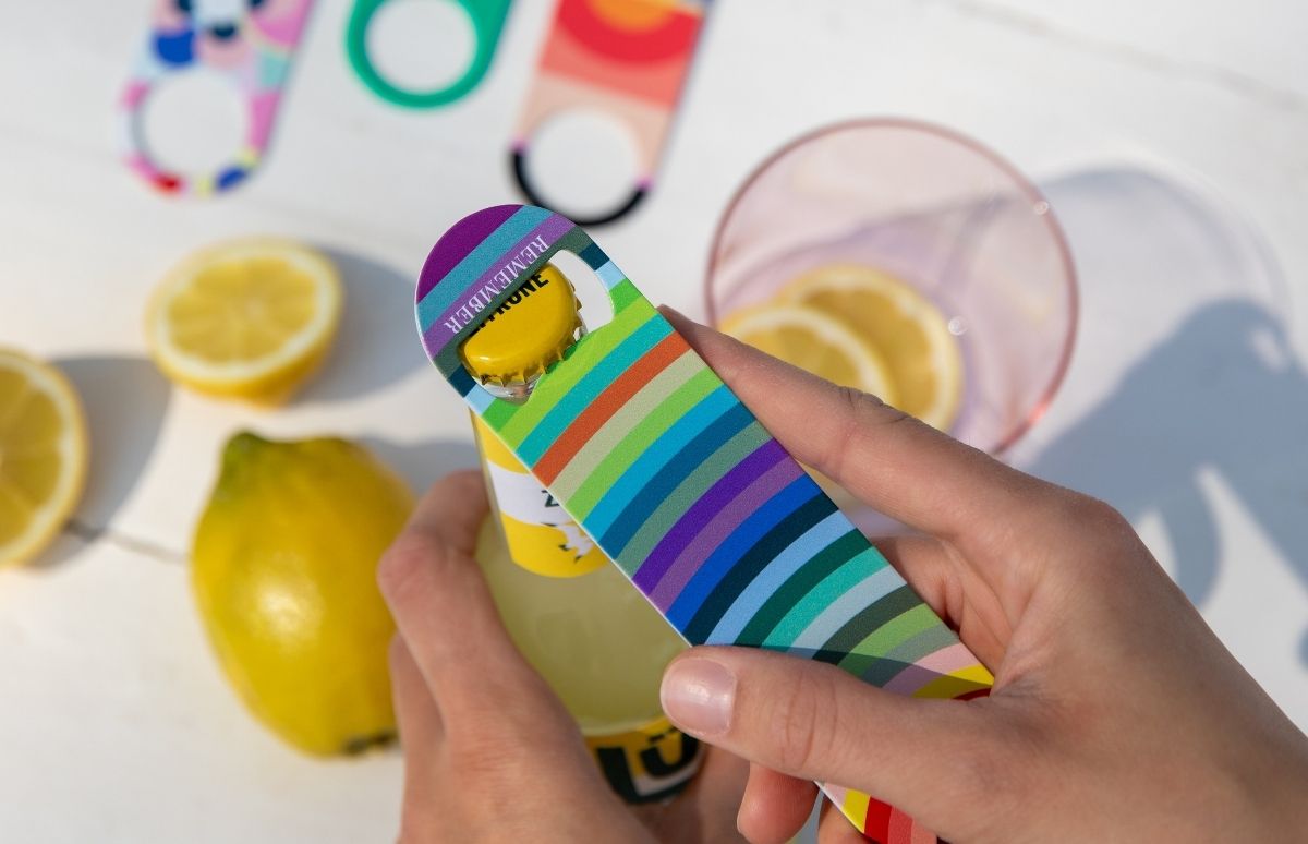 Zwei Hände öffnen eine gelbe Flasche mit einem bunten, gestreiften Flaschenöffner mit der Aufschrift „REMEMBER“. Im Hintergrund liegen frische Zitronen, ganze und halbe, sowie ein Glas mit Zitronenscheiben. Weitere Öffner unscharf.