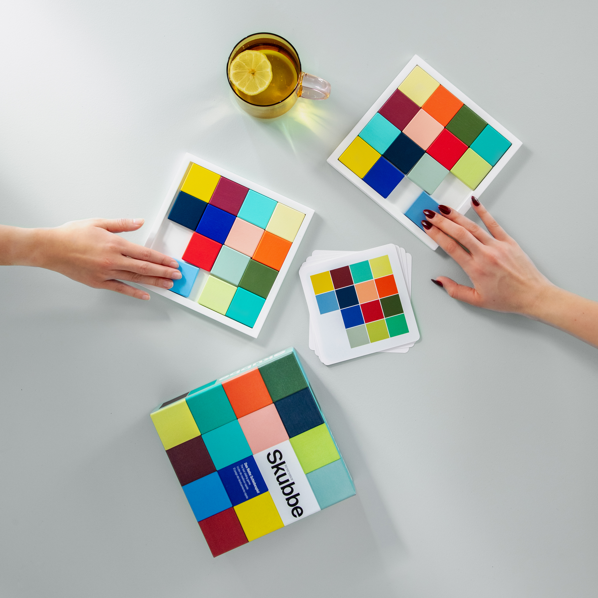 Vista superior del juego «SKUBBE»: dos manos mueven fichas de madera de colores sobre los tableros. También se ven las cartas del juego, el embalaje y una bebida.