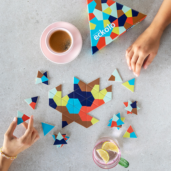 Zwei Hände legen bunte Dreiecks-Puzzleteile auf einen grauen Tisch. In der Mitte ein Muster. Daneben: rosa Kaffeetasse, Spielbox und Zitronenwasser.