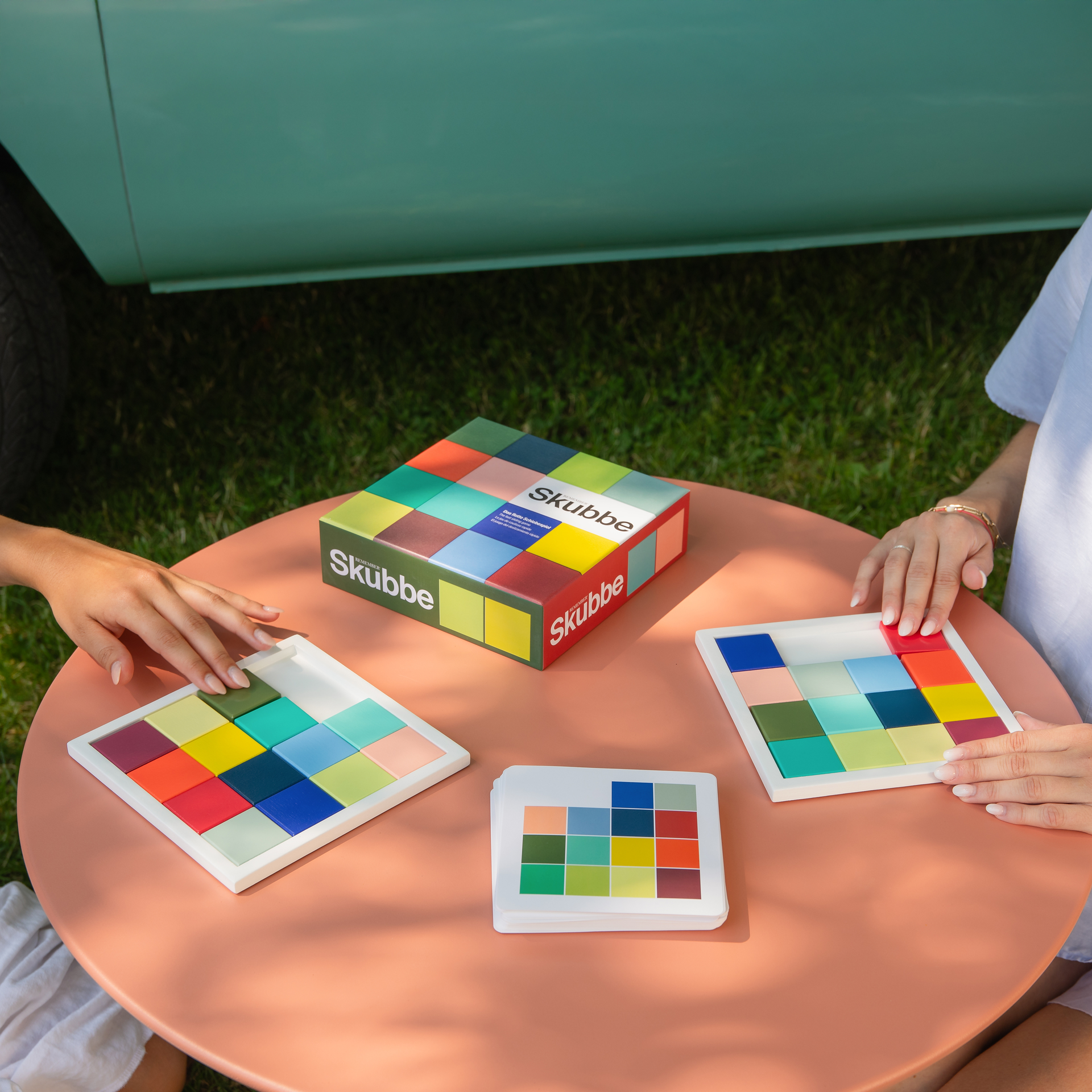 Dos personas juegan a Skubbe en una mesa. Se ven el tablero, las cartas y la caja. Hay fichas de colores sobre los tableros.