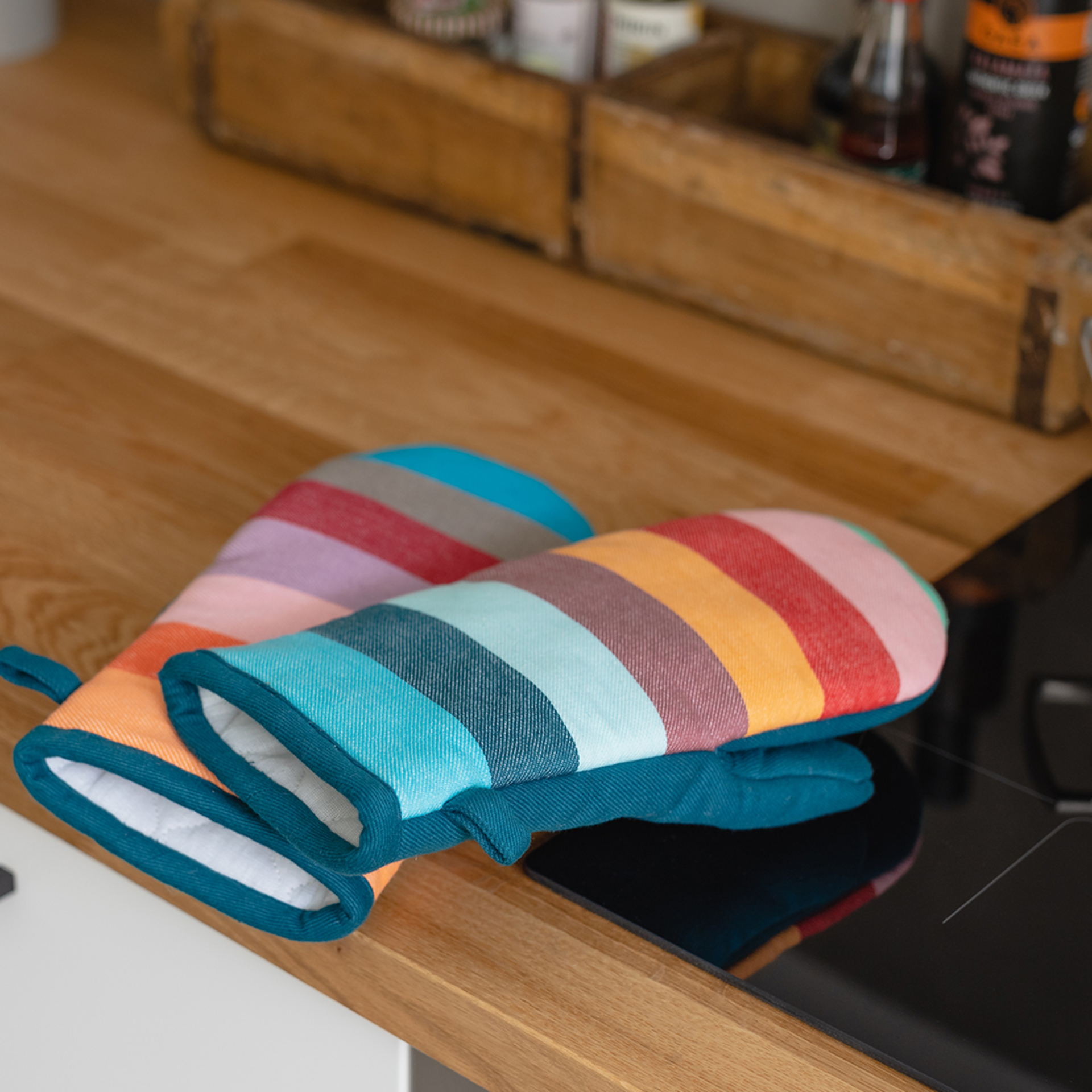 Due guanti da forno a righe in un set da 2 sono appoggiati l'uno sull'altro su una superficie di legno chiaro accanto a un piano cottura nero. Le strisce sono di colori diversi.