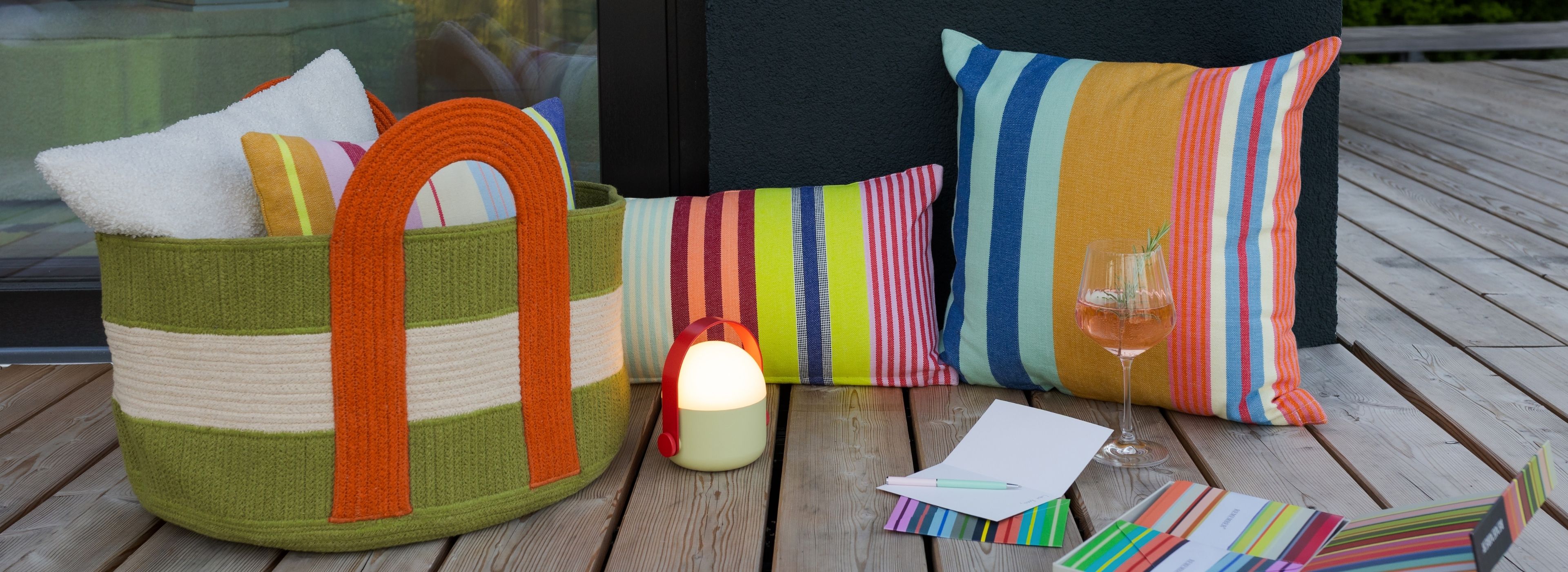 Grüne Korbtasche mit orangefarbenen Griffen auf Holzterrasse, gefüllt mit weißen Handtüchern und bunten Kissen. Daneben leuchtet eine kleine tragbare Lampe. Vorn: Rosé im Glas, Karte mit Stift und gestreifte Notizkarten.