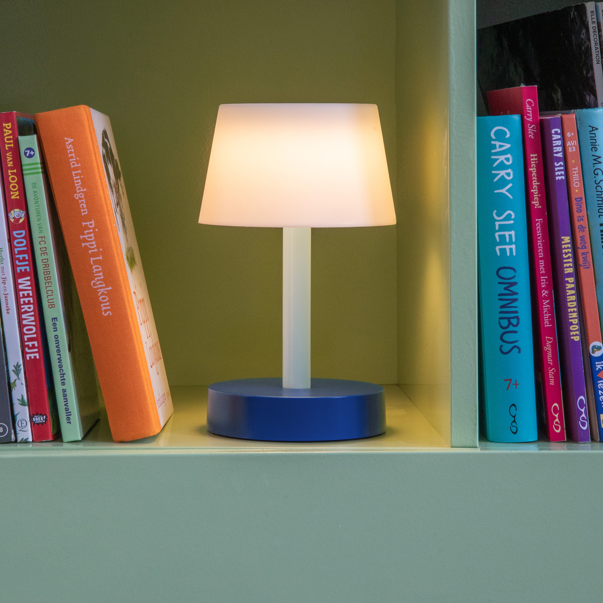 Sfeeropname van de Fritz 'sky' tafellamp met blauwe voet en lichtgevende witte kap op een plank tussen boeken.