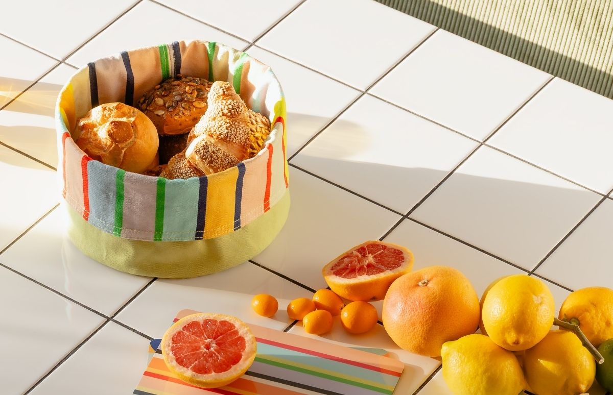 Bunter Brotkorb auf weißen Fliesen mit frischen Backwaren wie Brötchen, Kürbiskernbrot und Sesamcroissants. Daneben Zitrusfrüchte: rosa Grapefruits, Kumquats und gelbe Zitronen. Sonnenlicht sorgt für Morgenstimmung.