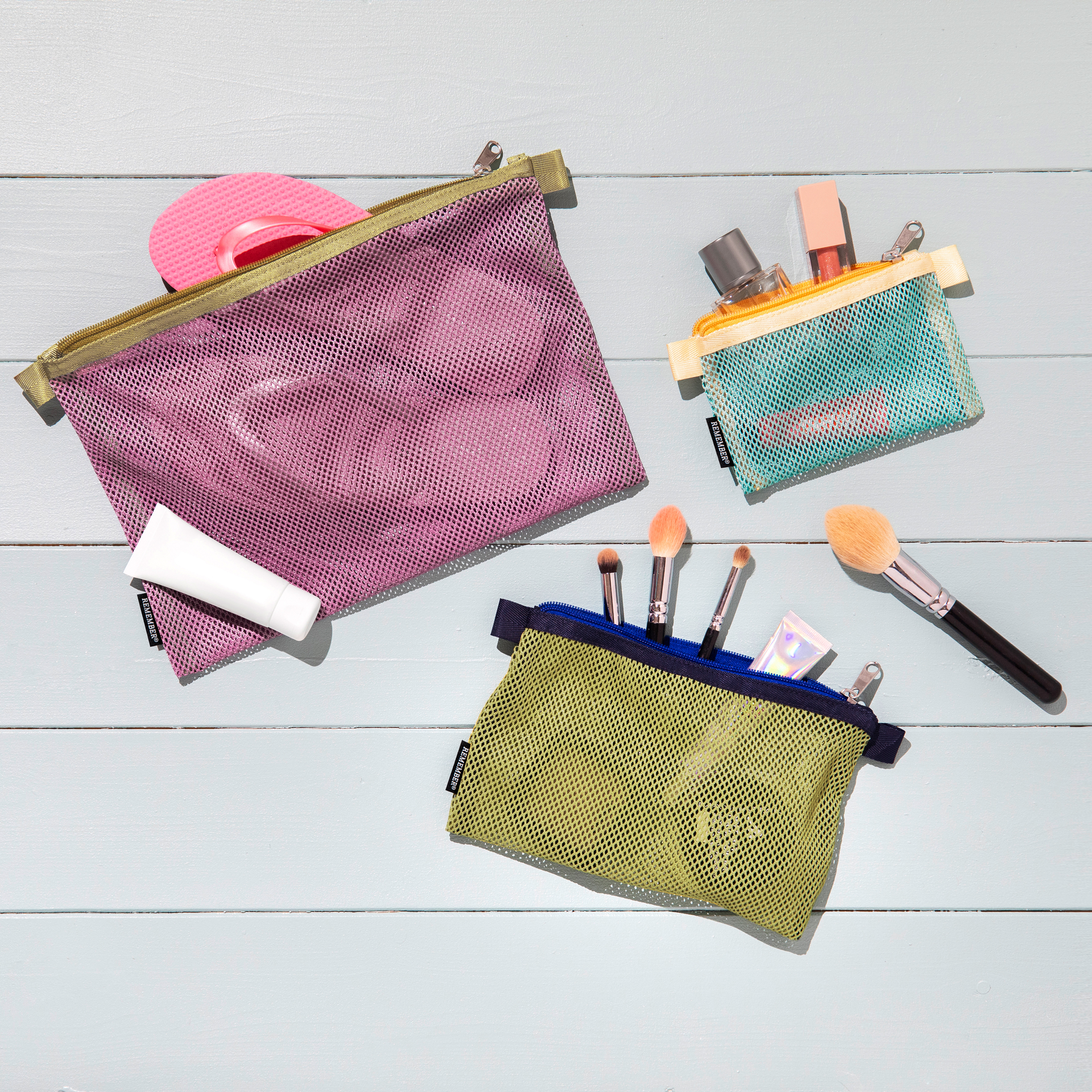 Three coloured mesh bag 'Jordy' sets, filled with cosmetics, on a light blue wooden background. The bags are in pink, turquoise and olive green.
