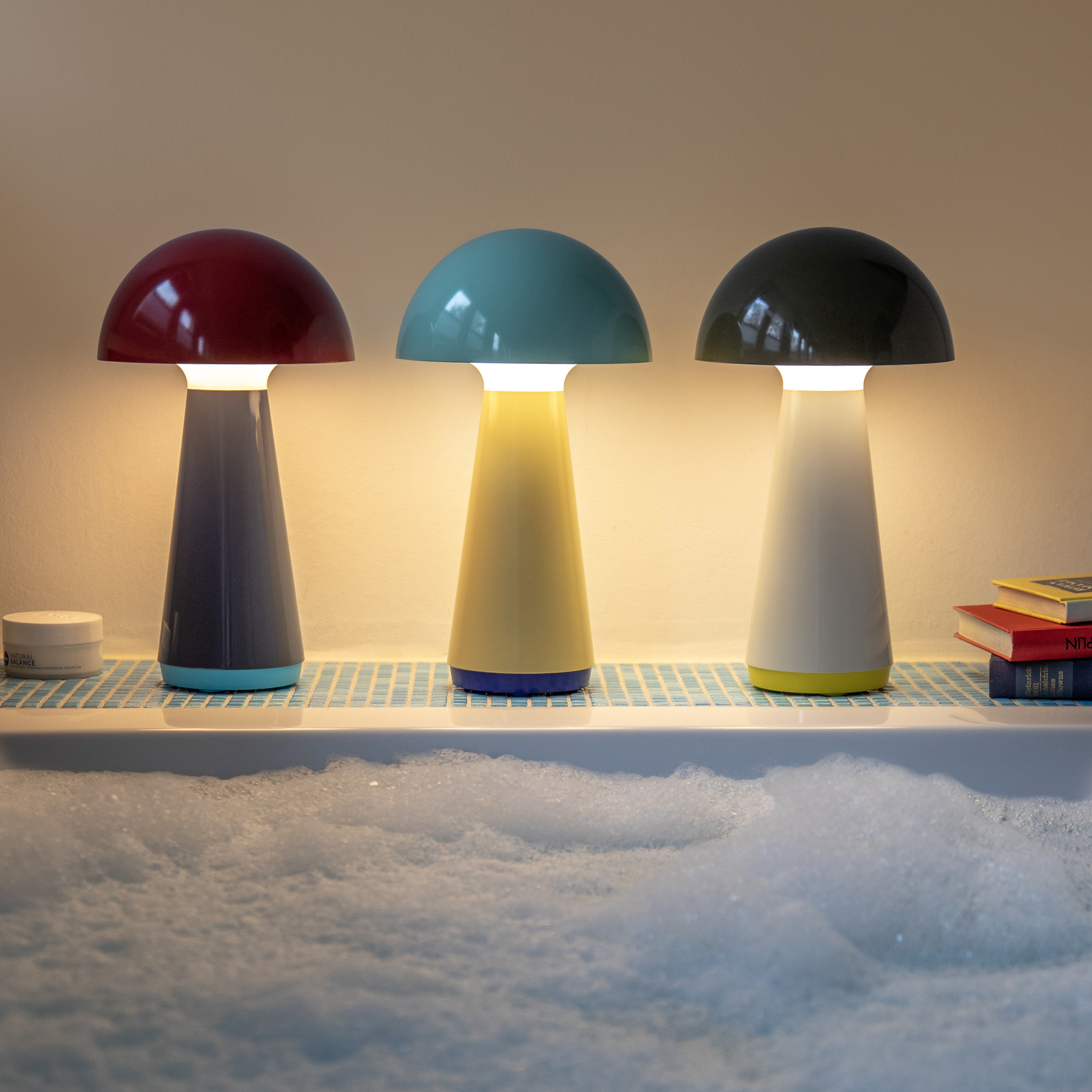 Drie 'Bob' tafellampen in rood, mint en olijfgroen. Ze staan op een blauw mozaïek tafeltje naast een bad met schuim. Elke lamp heeft een warme witte gloed.