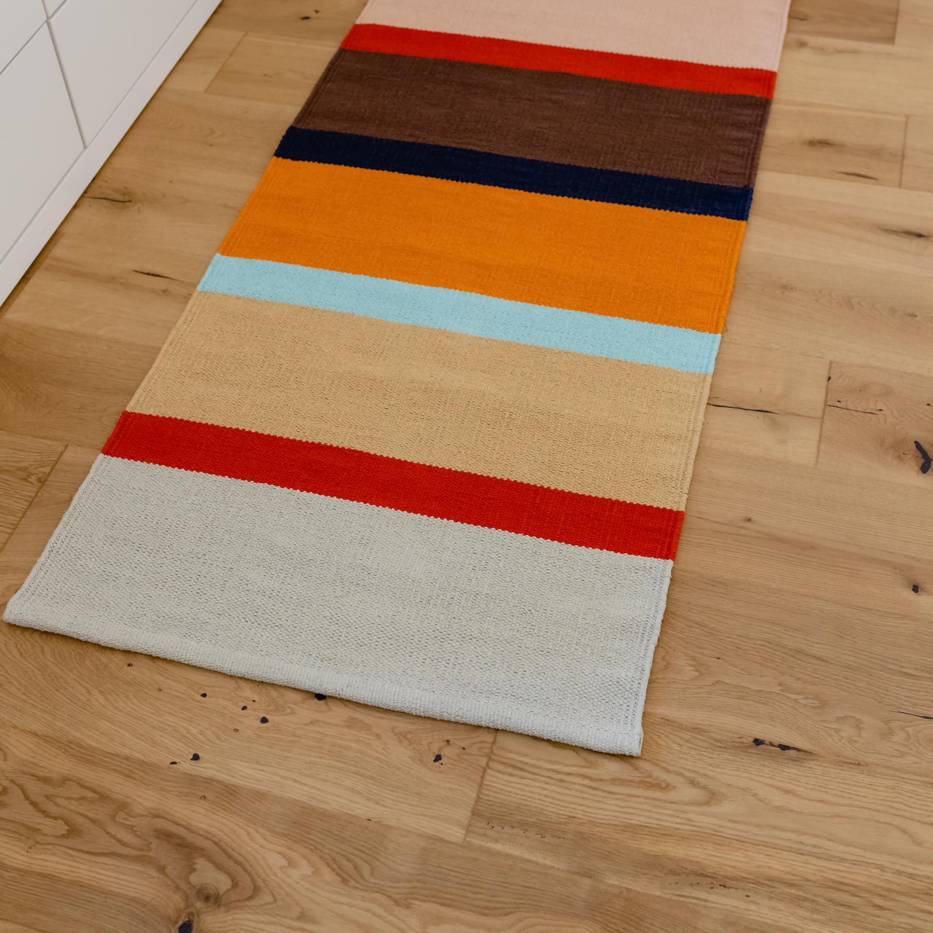 Lang gestreifter Teppichläufer 'Zaza' auf Holzboden. Die Streifen sind in Farben wie Rot, Orange, Braun, Beige, Hellblau und Grau gehalten.