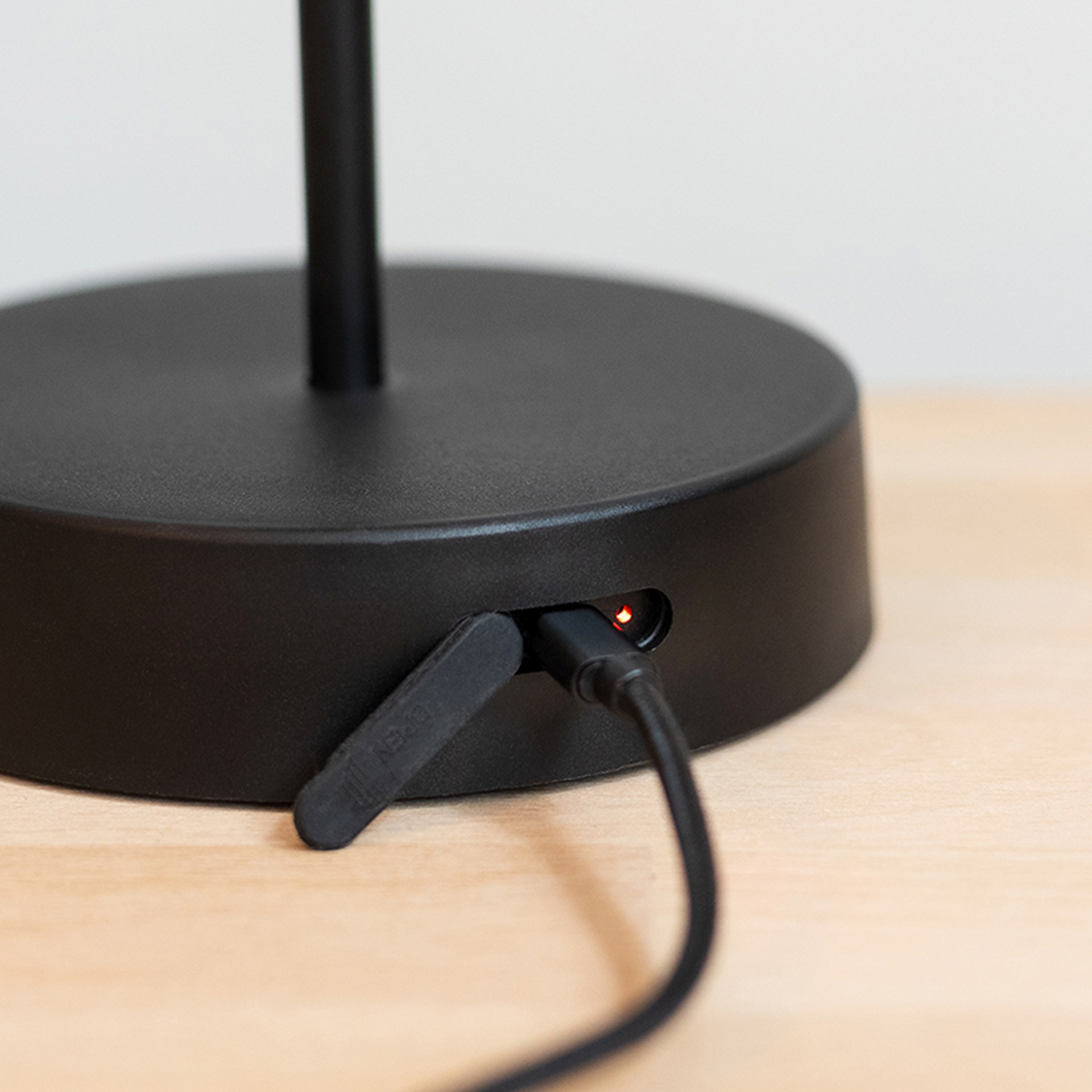 Close-up of the Oscar table lamp in 'pure' design with plugged-in USB cable and red charging indicator on the base on a light-coloured table.