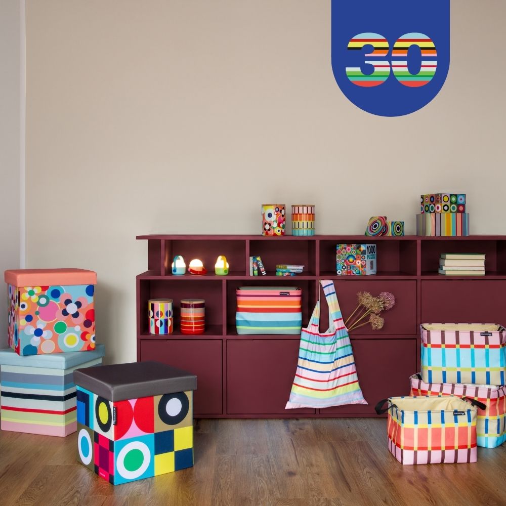 Farbenfroher Raum mit bordeauxrotem Regal, gefüllt mit bunten Aufbewahrungsboxen und Dekoartikeln. Auf dem Holzboden stehen gemusterte Hocker und Körbe. Oben rechts an der Wand ist ein blaues Logo mit der Zahl „30“.