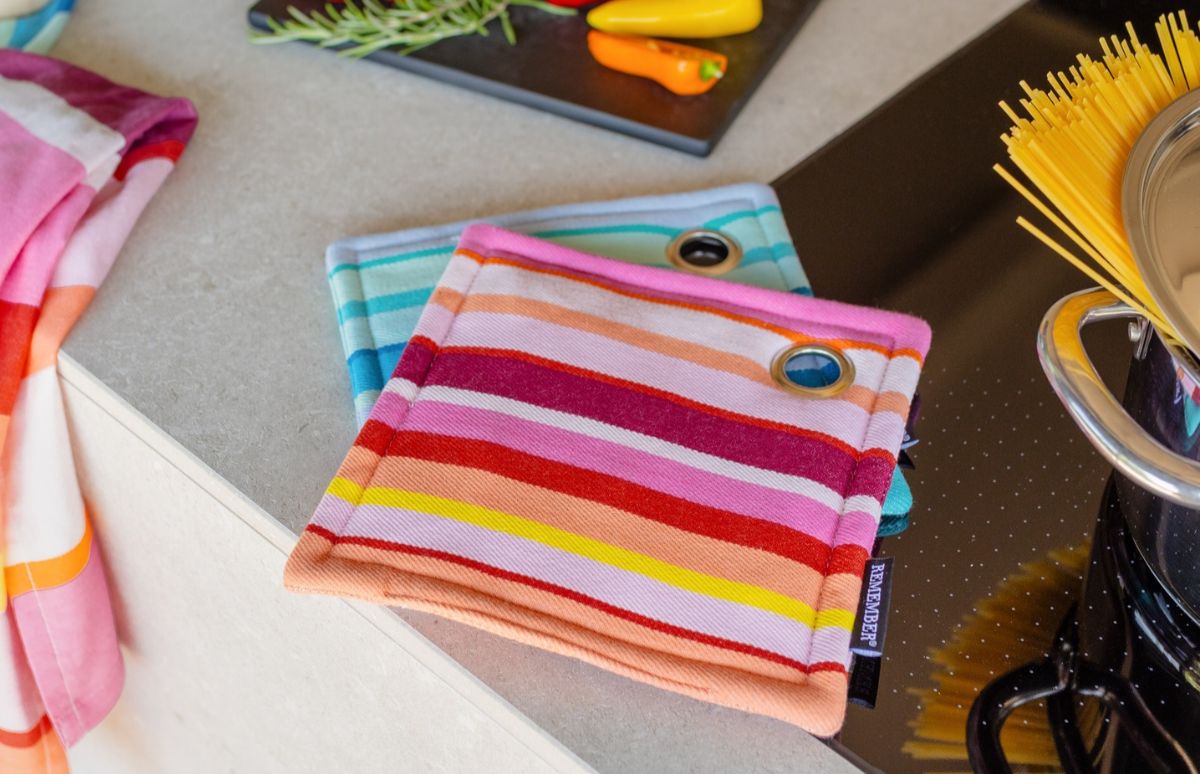 Zwei bunte, gestreifte Topflappen mit Metallösen liegen auf einer hellgrauen Küchenarbeitsplatte. Oben Streifen in Rosa, Rot, Orange und Gelb, unten in Türkis und Hellblau. Links ein passendes Küchenhandtuch.