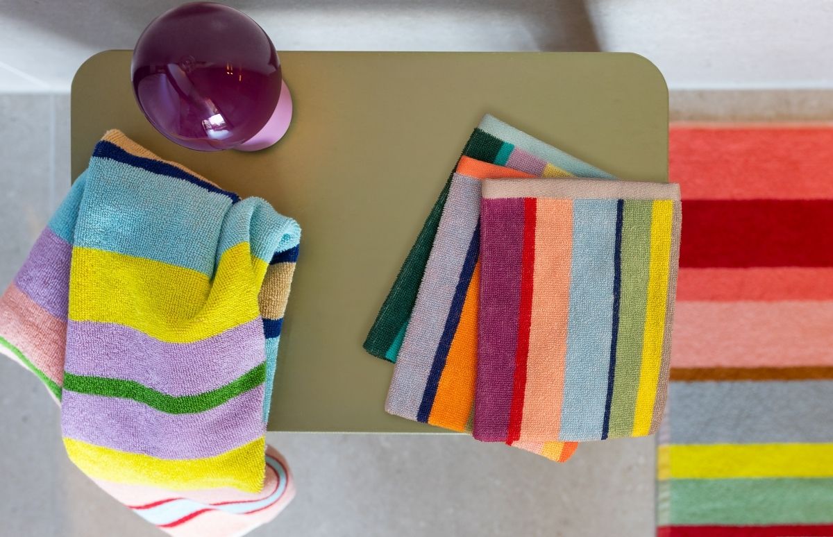 Lebhaft gestreifte Handtücher in Blau, Gelb, Lila und Grün liegen auf einer olivgrünen Ablage. Oben links steht eine glänzende violette Kugellampe. Rechts ist ein mehrfarbig gestreifter Teppich zu sehen.