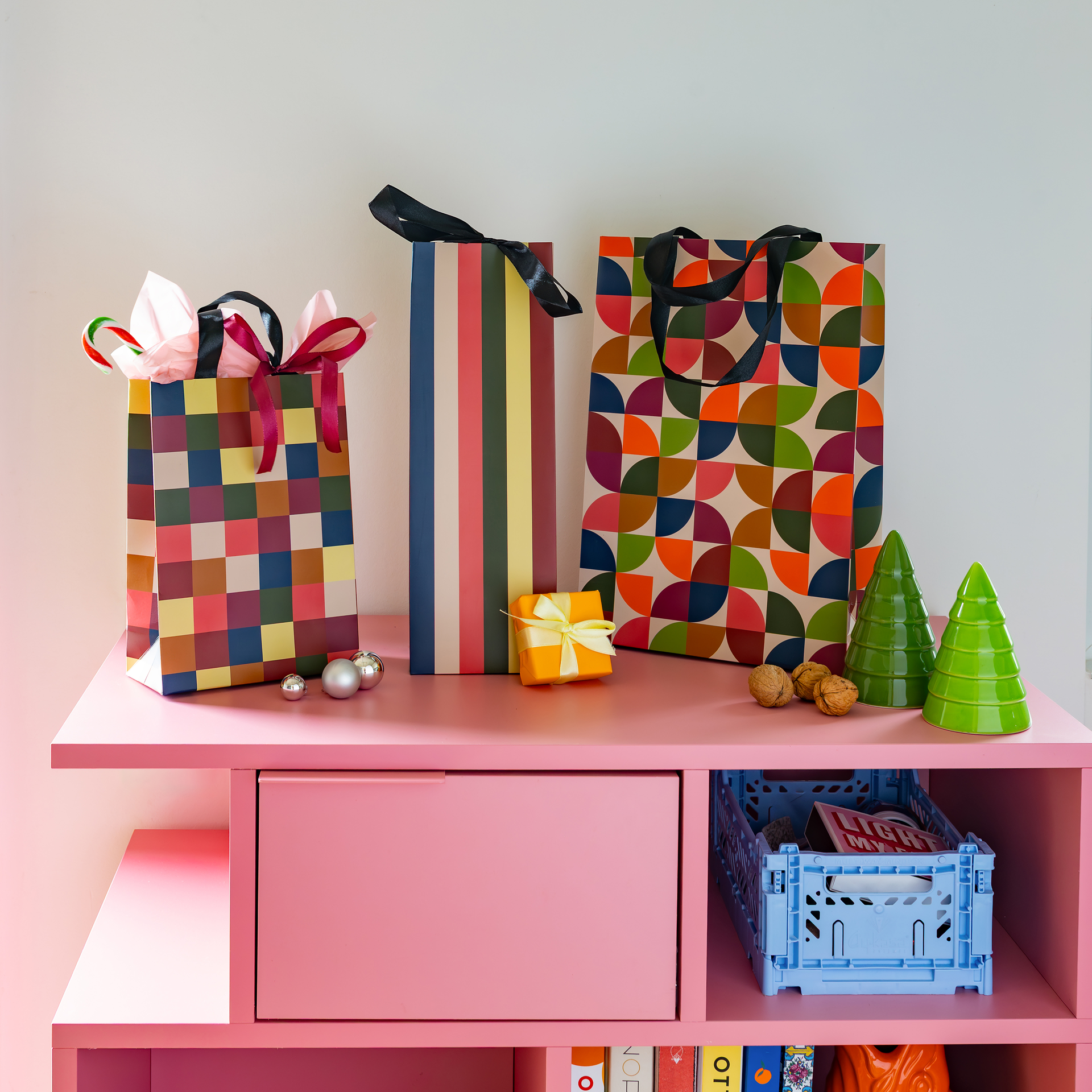 Tres bolsas de regalo de colores con estampados y cintas sobre una estantería rosa. Un pequeño regalo amarillo, abetos verdes, nueces y bolas plateadas completan la escena festiva. Una atractiva presentación de envoltorios para regalos.