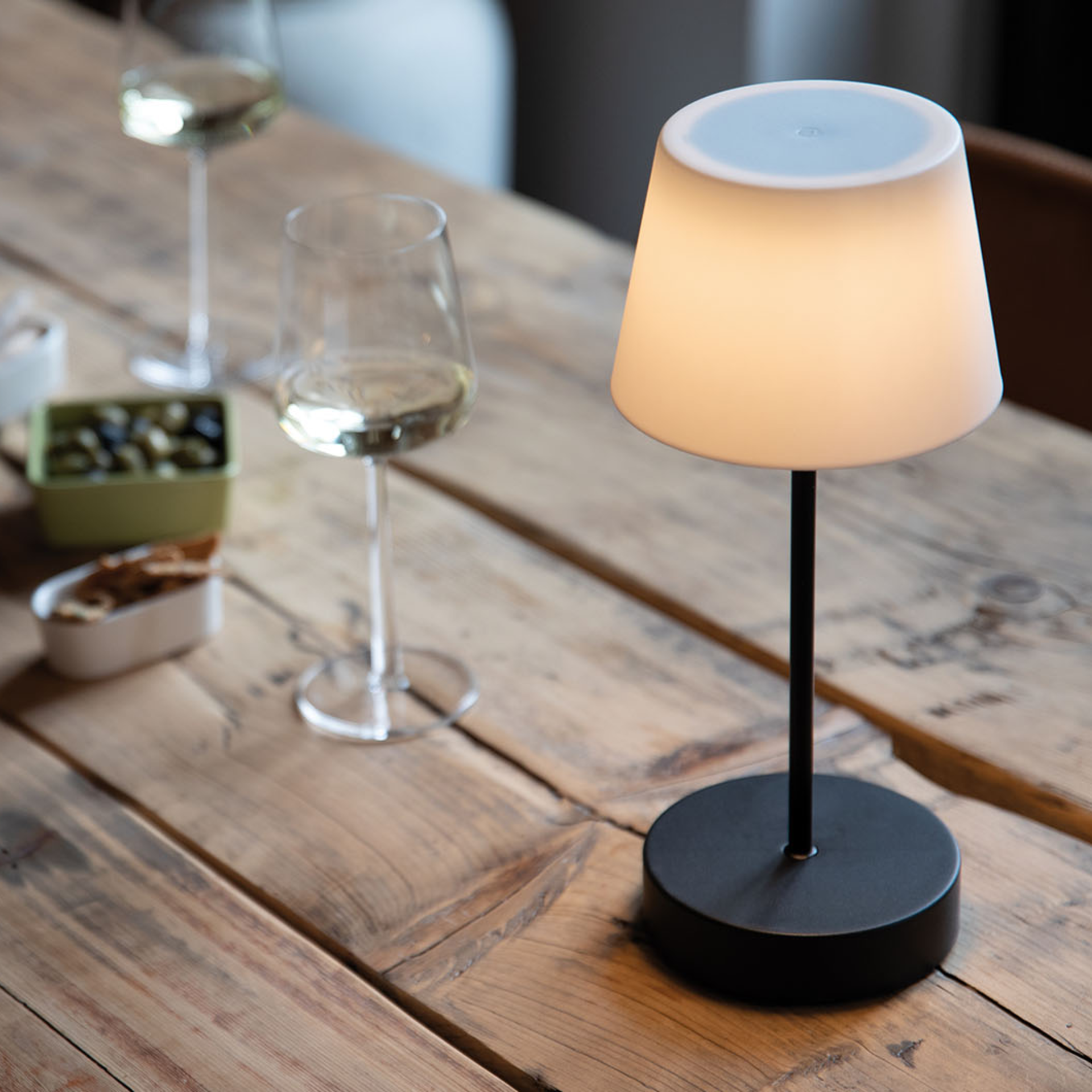 The Oscar 'pure' table lamp on a wooden table. The lamp has a black base and a luminous white lampshade. Glasses and bowls with food can be seen in the background.