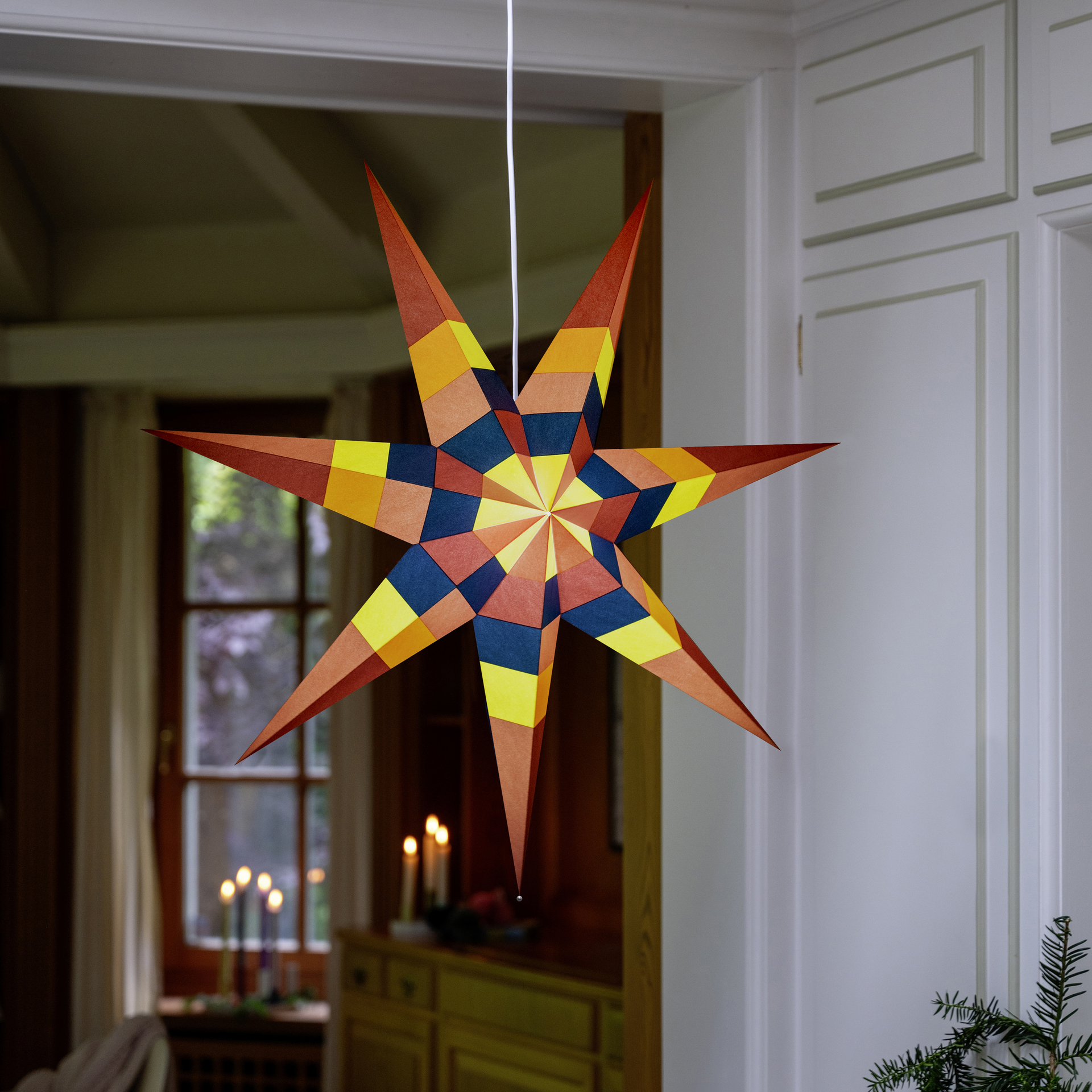 Farbenfroher Papierstern mit sieben zackigen Spitzen, Oberfläche aus vielen gefalteten geometrischen Flächen in Rot, Orange, Gelb, Dunkelblau und Rosa. Radiales Muster um die Mitte. Oben weiße Kordel, unten silberner Stopper.