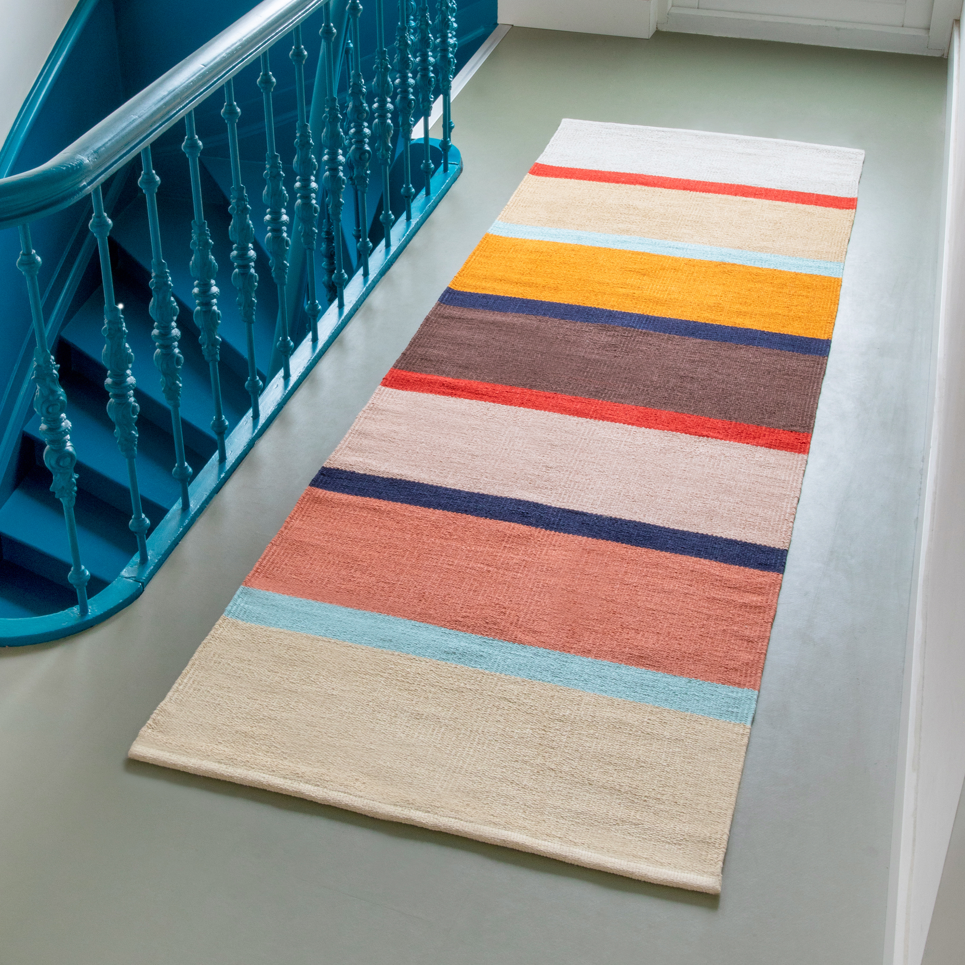 Handgewebter Teppichläufer 'Zaza' mit Streifenmuster in Beige, Blau, Orange, Braun und Rot auf einem grauen Boden neben einer blauen Treppe.