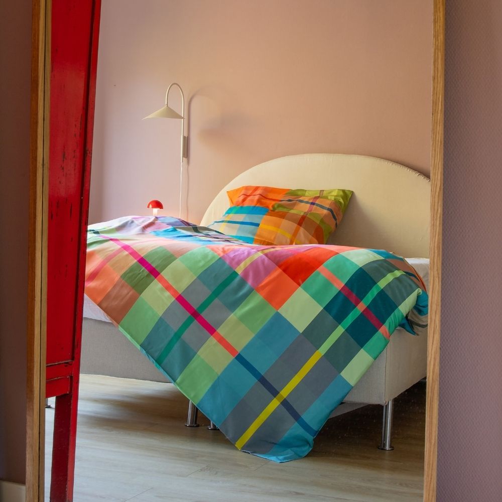 Ein gemütliches Schlafzimmer, im Spiegel reflektiert. Das Bett mit hellem Kopfteil ist mit farbenfroher, karierter Bettwäsche in Blau, Grün, Orange, Pink und Gelb bezogen. Eine weiße Wandleuchte und eine kleine rote Pilzlampe schmücken die zartrosa Wand.