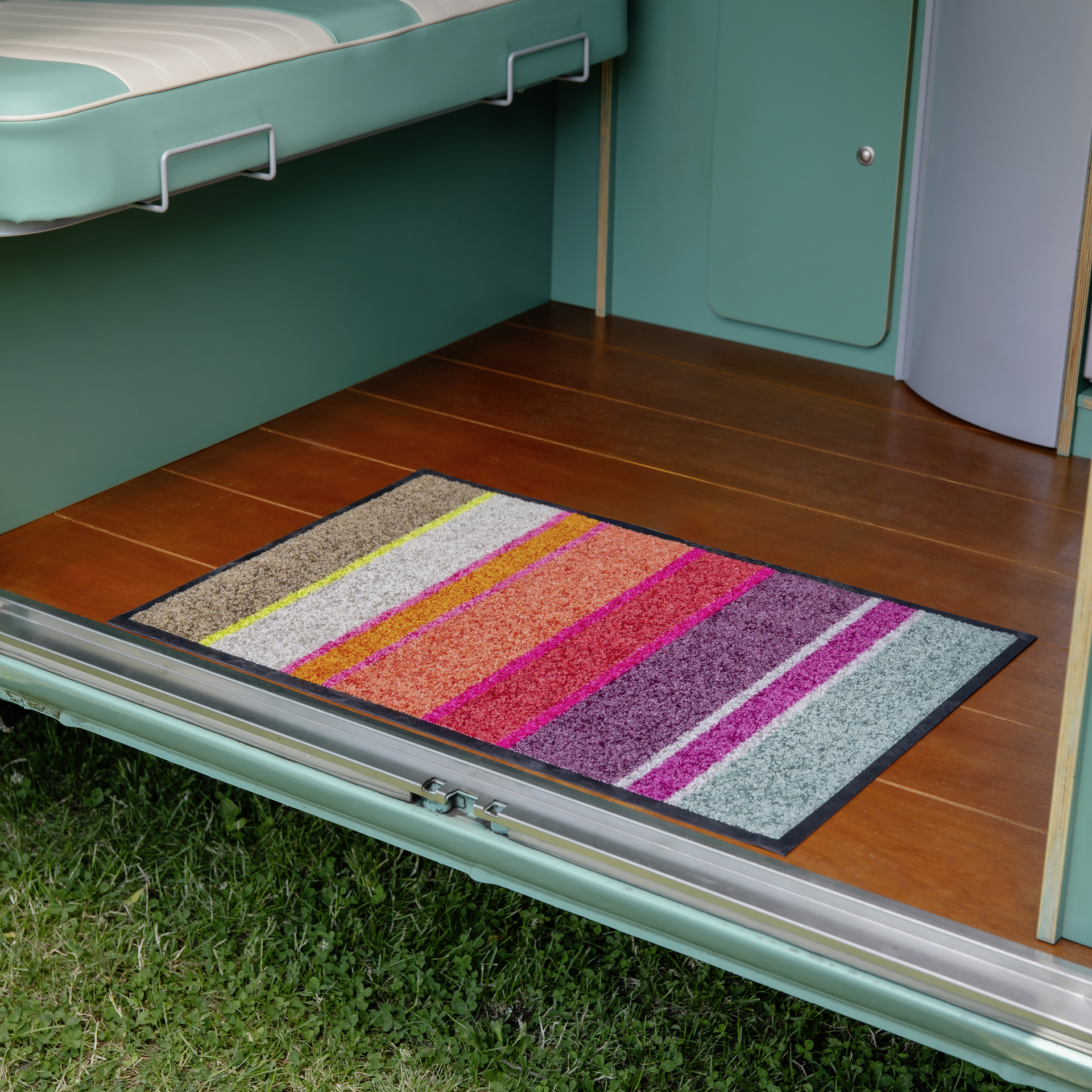 Farbige Fußmatte 'Largo' mit Streifenmuster in Braun, Gelb, Orange, Rosa, Lila und Grau auf Holzboden vor einer grünen Wand.