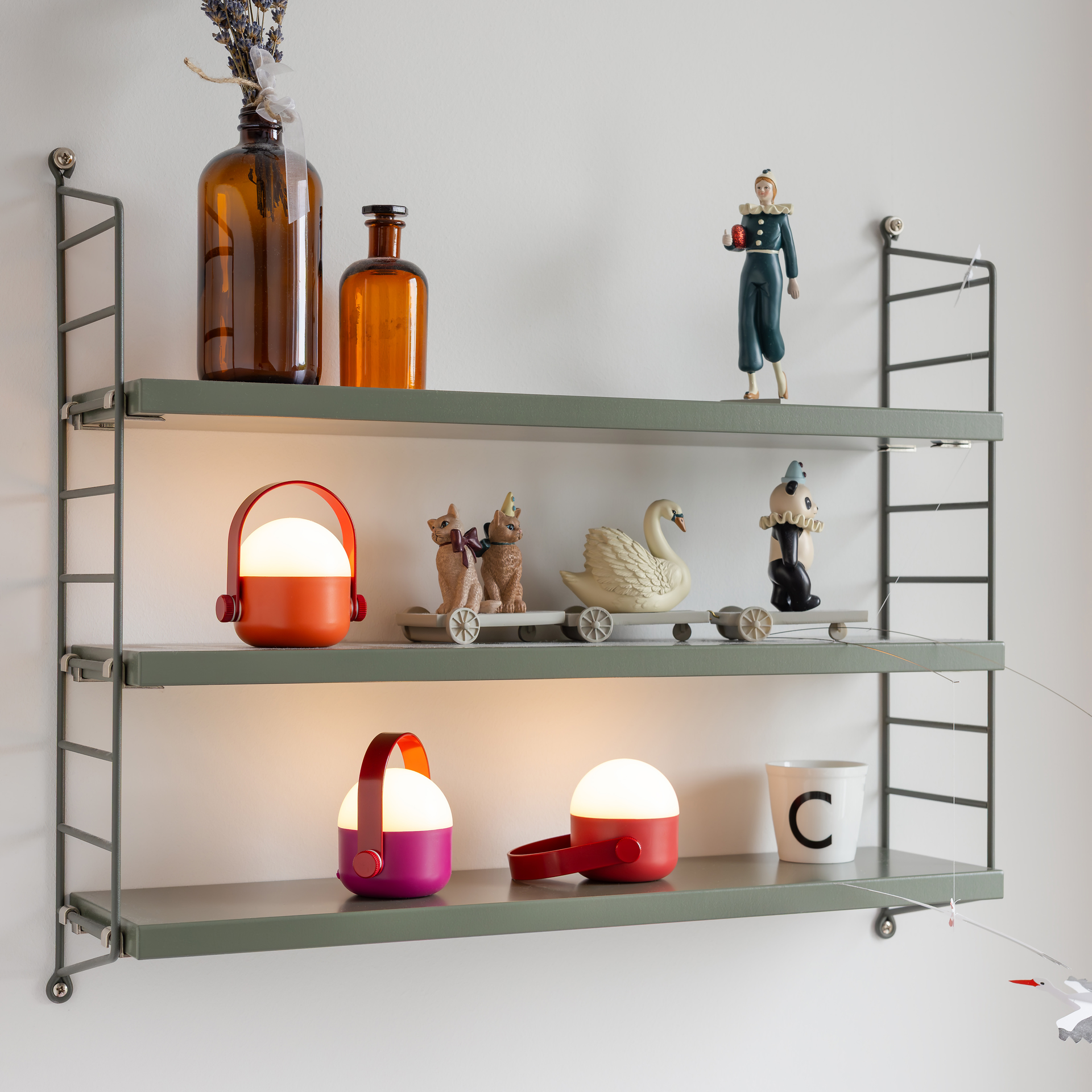 Drei OLE mini LED-Leuchten, rot-orange, purpurrot und rot, eines 3er-Sets leuchten warmweiß auf grünen Regalen. Sie sind umgeben von braunen Flaschen und Figuren wie Katzen und einem Schwan und erzeugen eine gemütliche Akzentbeleuchtung.