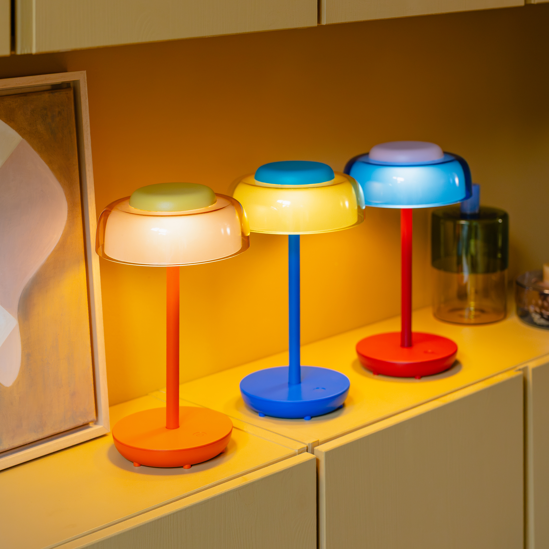 Drie Juno-tafellampen in verschillende felle kleuren op een plank. Gekleurde stelen en heldere lampenkappen stralen in warme gele tinten en koel blauw. De set toont de diversiteit van de draadloze Juno-serie.