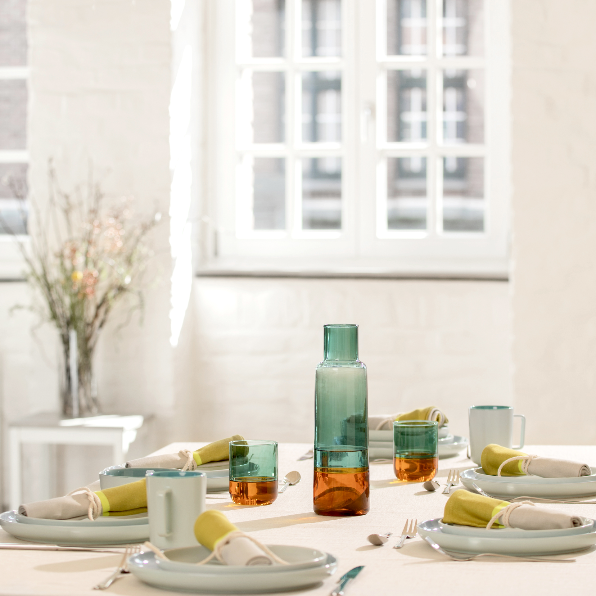 A table setting with plates, napkins, cutlery, cups and glasses 'amber' in a set of 2. A glass carafe is placed in the centre. Bright daylight in the background.
