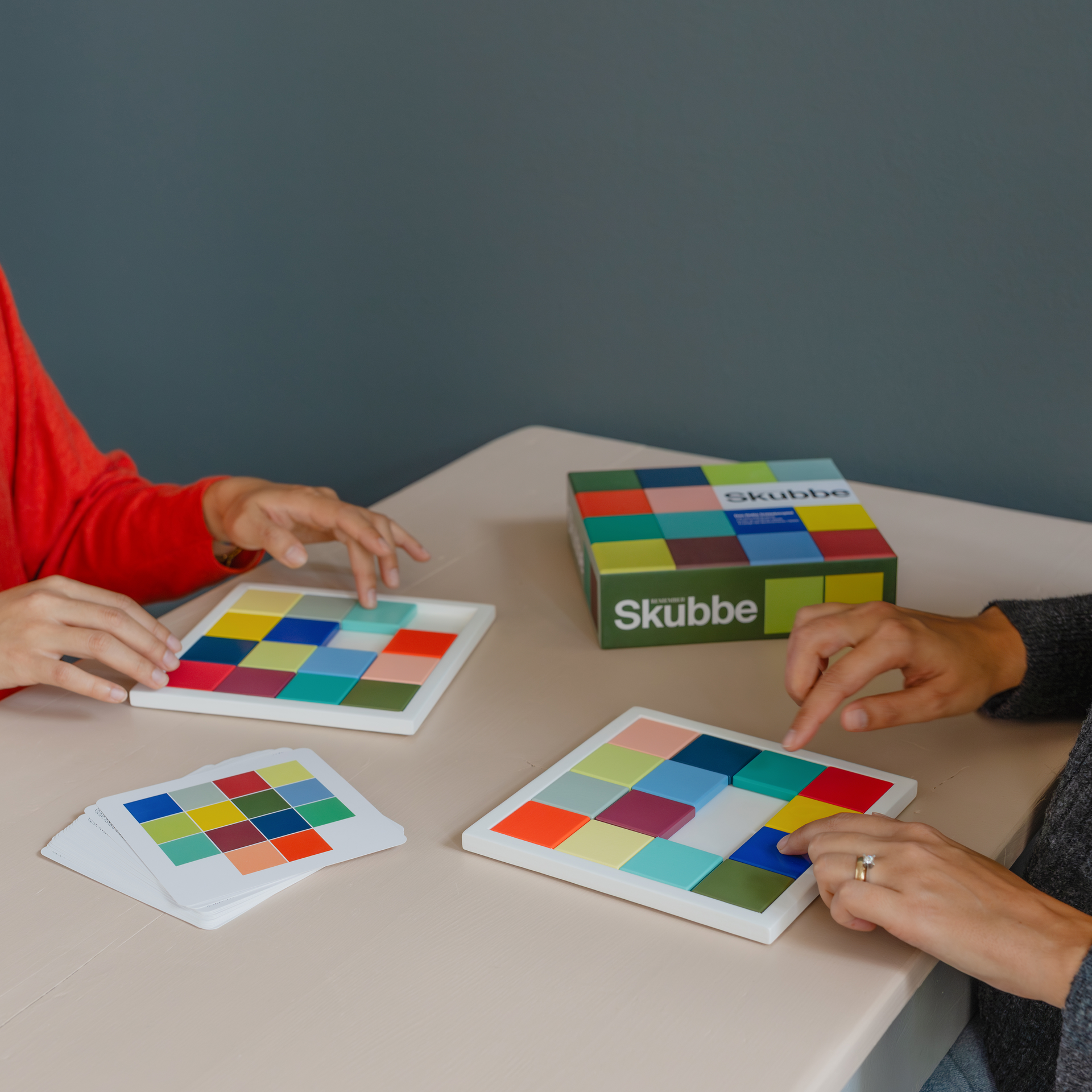 Dos personas juegan a «SKUBBE»: fichas de madera de colores en un tablero de juego con cartas de referencia y estuche.