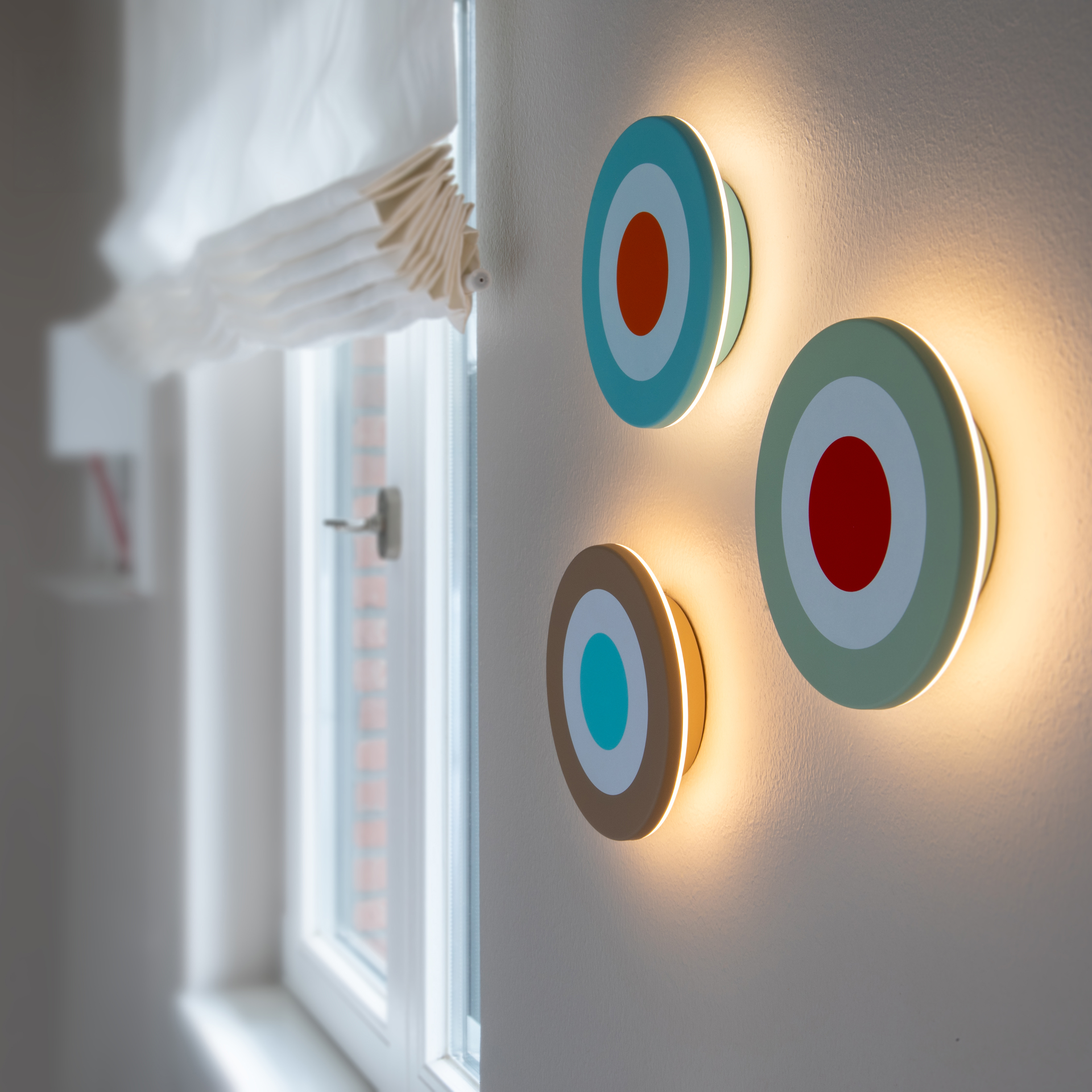 Runde Wandleuchte mit modernem, grafischem Kreisdesign in Mintgrün, Weiß und Rot, schräg von rechts fotografiert. Zentrales rotes Dimmfeld, umgeben von weißem und mintgrünem Ring. Akkubetriebene LED mit warmweißem Licht.