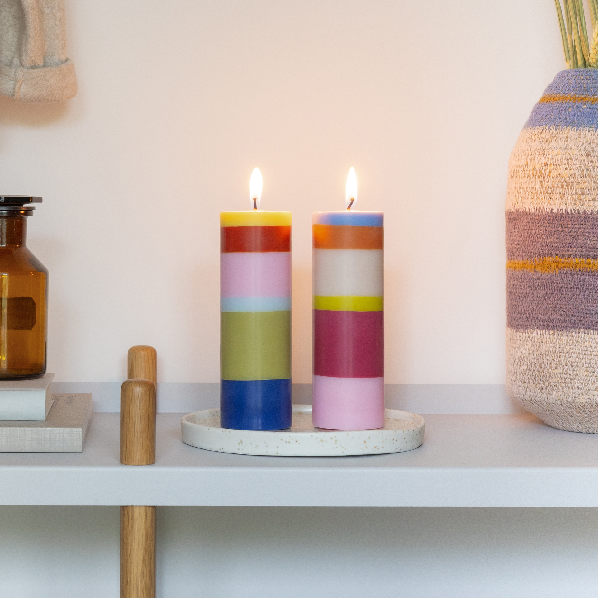Zwei hohe Stumpenkerzen im Set, je sechs horizontale Farbschichten. Links: Limettengrün, Rot, Hellrosa, Hellblau, Olivgrün, Dunkelblau. Rechts: Hellblau, Orange, Beige, Gelb, Magenta, Hellrosa. Weißer Docht oben.