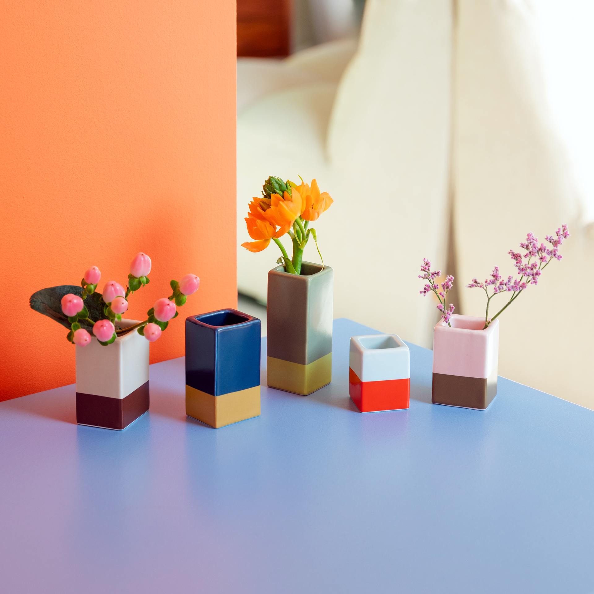Fünf farbige Vasen mit Blumen auf einem hellblauen Tisch, vor einer Wand in Orange und einem hellen Hintergrund. Die Vasen sind unterschiedlich groß.