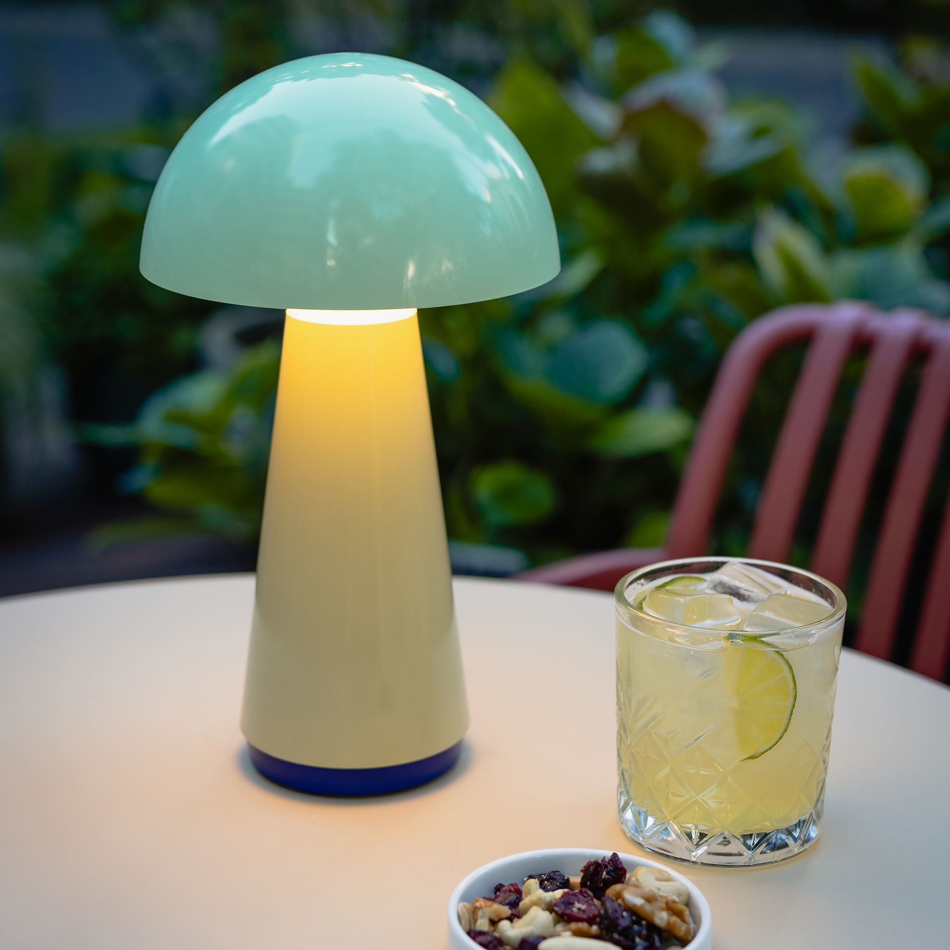 La lámpara de mesa color menta «Bob» ilumina una mesa, junto a un cóctel y un cuenco con frutos secos. Al fondo se ven plantas verdes y una silla.