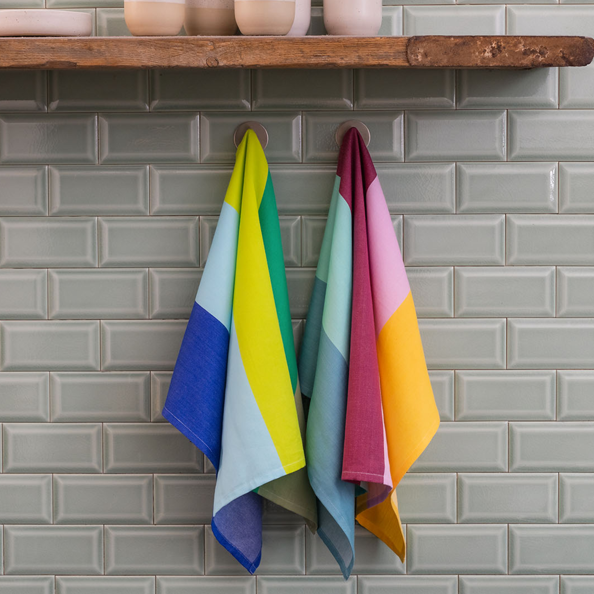 Deux sets de lingettes séchantes multicolores sont suspendus à des crochets devant un mur de carrelage vert clair. Au-dessus des serviettes se trouve une étagère en bois avec de la vaisselle.